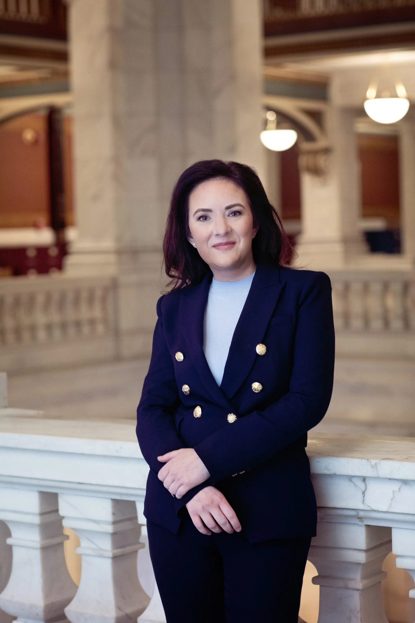 A woman with dark shoulder-length hair, dressed in a navy blazer with gold buttons, stands in front of a marble and wood interior with warm lighting, looking at the camera.