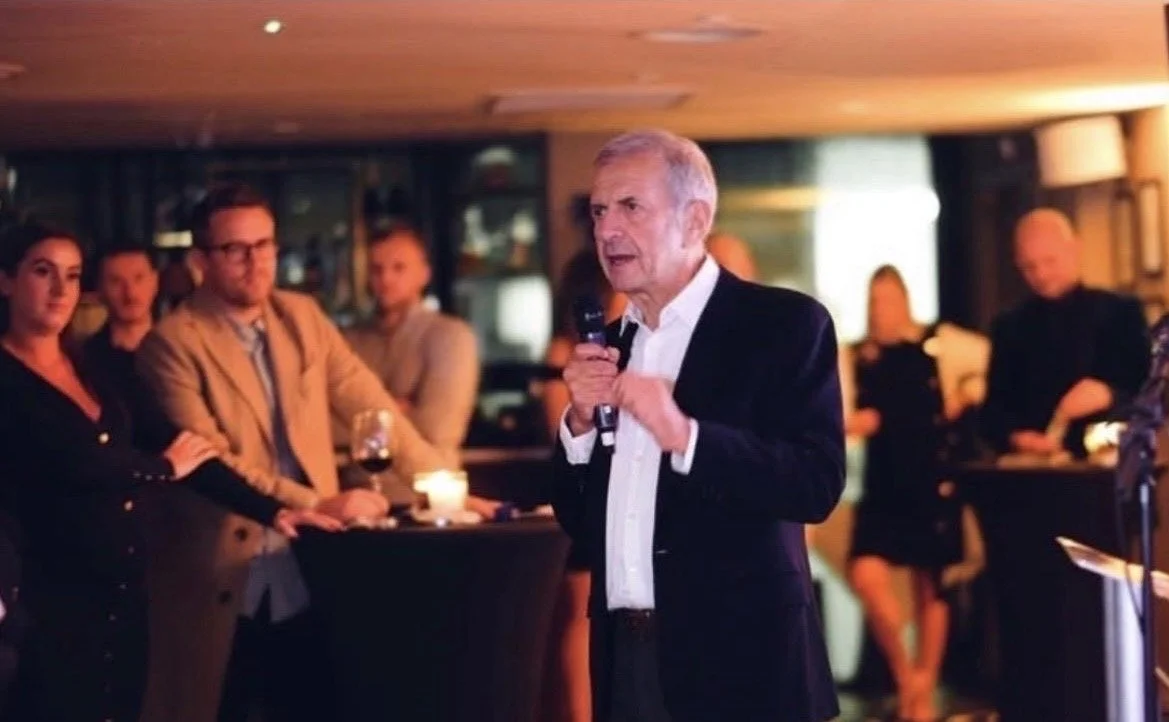 An older man in a black suit and white shirt speaking into a microphone at an indoor event, with an audience of casually dressed people watching him.