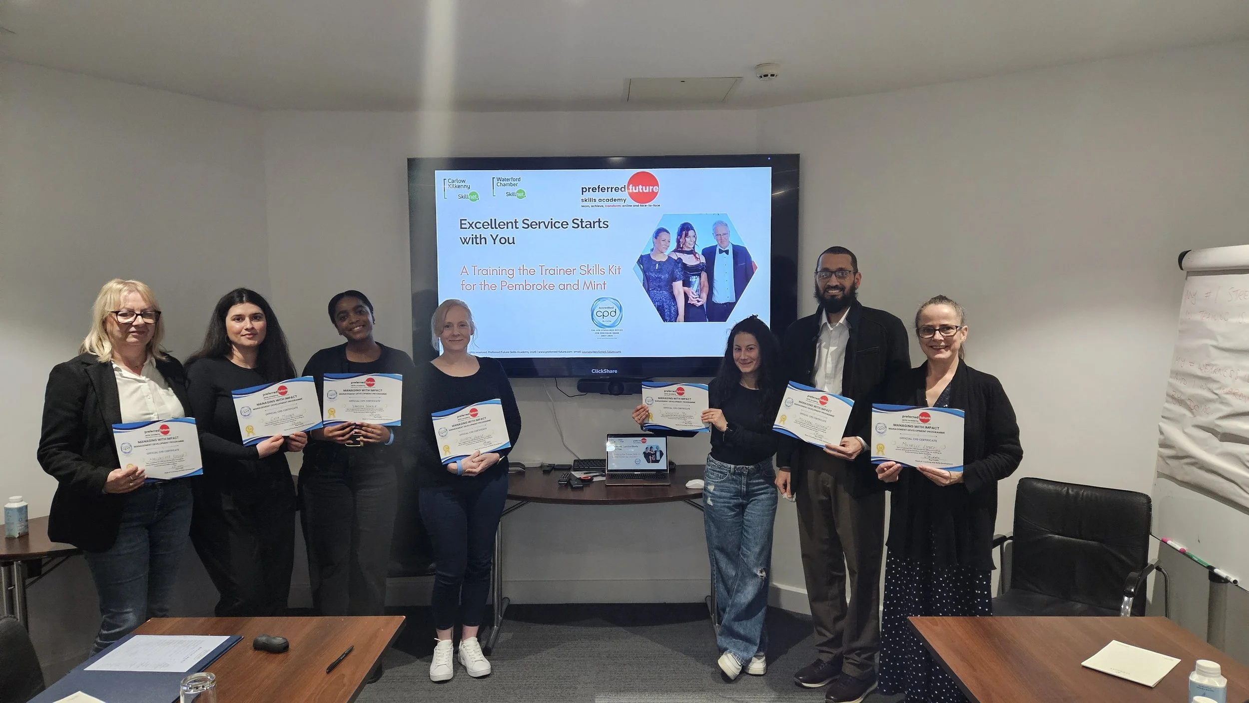 Group of seven people standing in a conference room, holding certificates, with a presentation screen behind them displaying a training event for the Preferred Future Skills Academy.