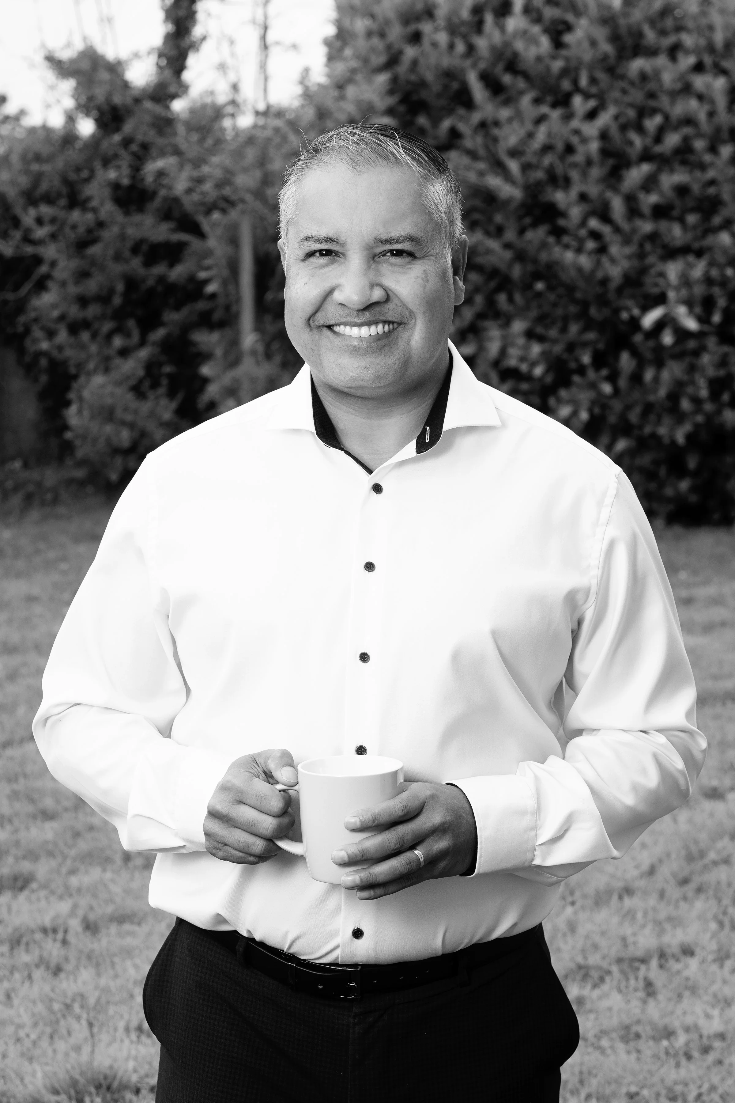 A man smiling, standing outdoors with trees and bushes in the background, holding a white mug in both hands.