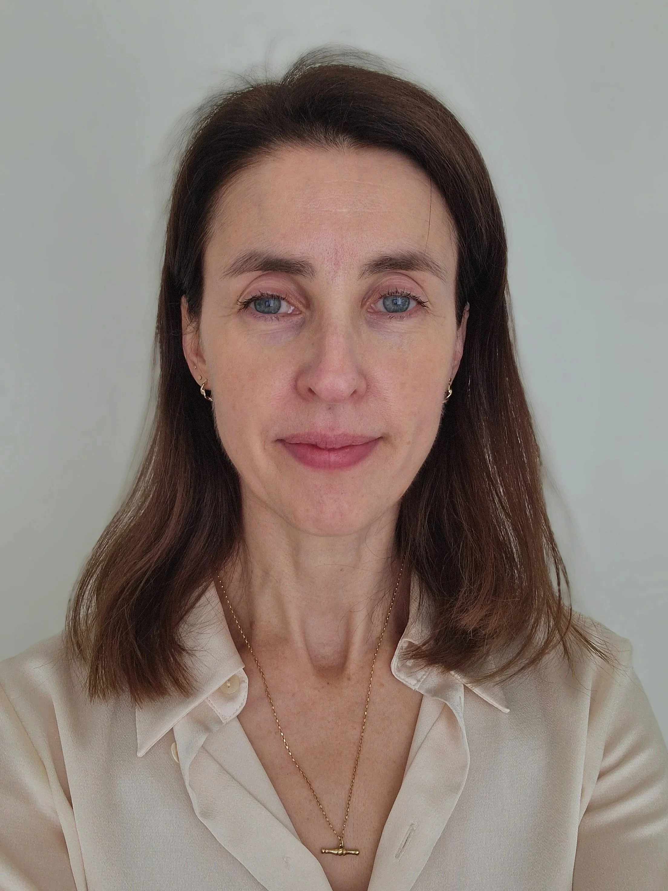A woman with brown hair, wearing a beige blouse, gold jewelry including earrings and a necklace with a pendant, standing against a plain, light-colored background.