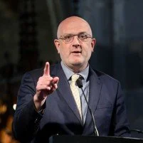 A man with glasses speaking at a podium in a suit, gesturing with his right hand.