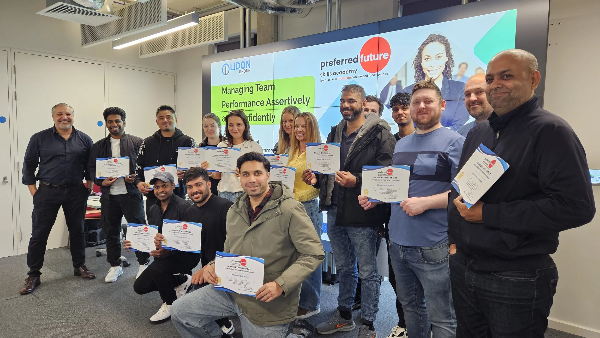 Group of people at a certificate award ceremony, holding certificates, in a conference room with a colorful presentation screen in the background.