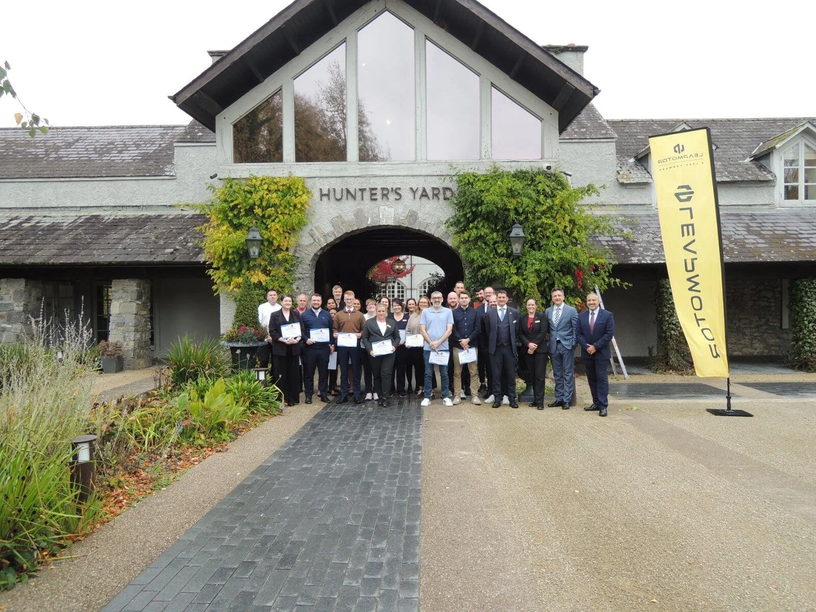 Group of people standing in front of a building with a sign that reads 'Hunter's Yard', some holding certificates, next to a yellow banner with 'EFTOMC' and other text.