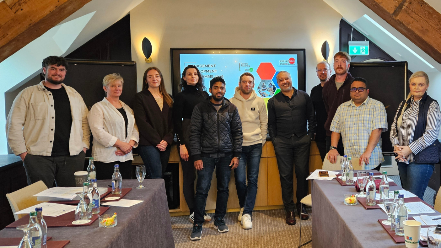 Group of twelve people standing in a conference room with tables and bottled water, in front of a large screen displaying a presentation slide titled 'Management Development Programme 2023'.