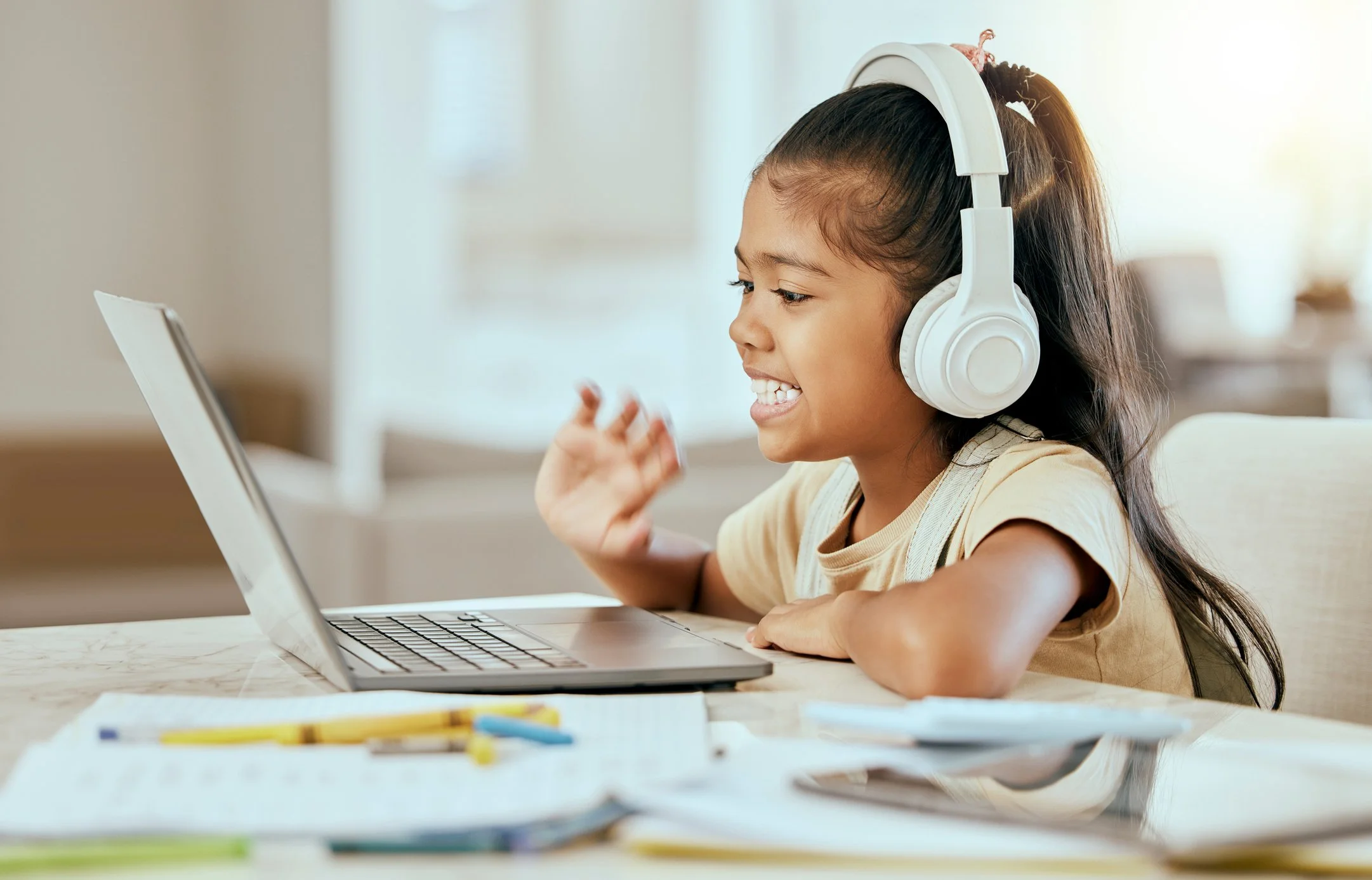 A young girl with long dark hair wearing a beige shirt, smiling, and wearing large white headphones, engaging with a laptop at a white table with papers and colored pencils.