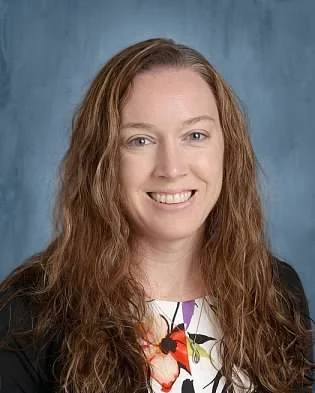Portrait of a smiling woman with long wavy red hair, wearing a black blazer and a white floral blouse, against a blue background.