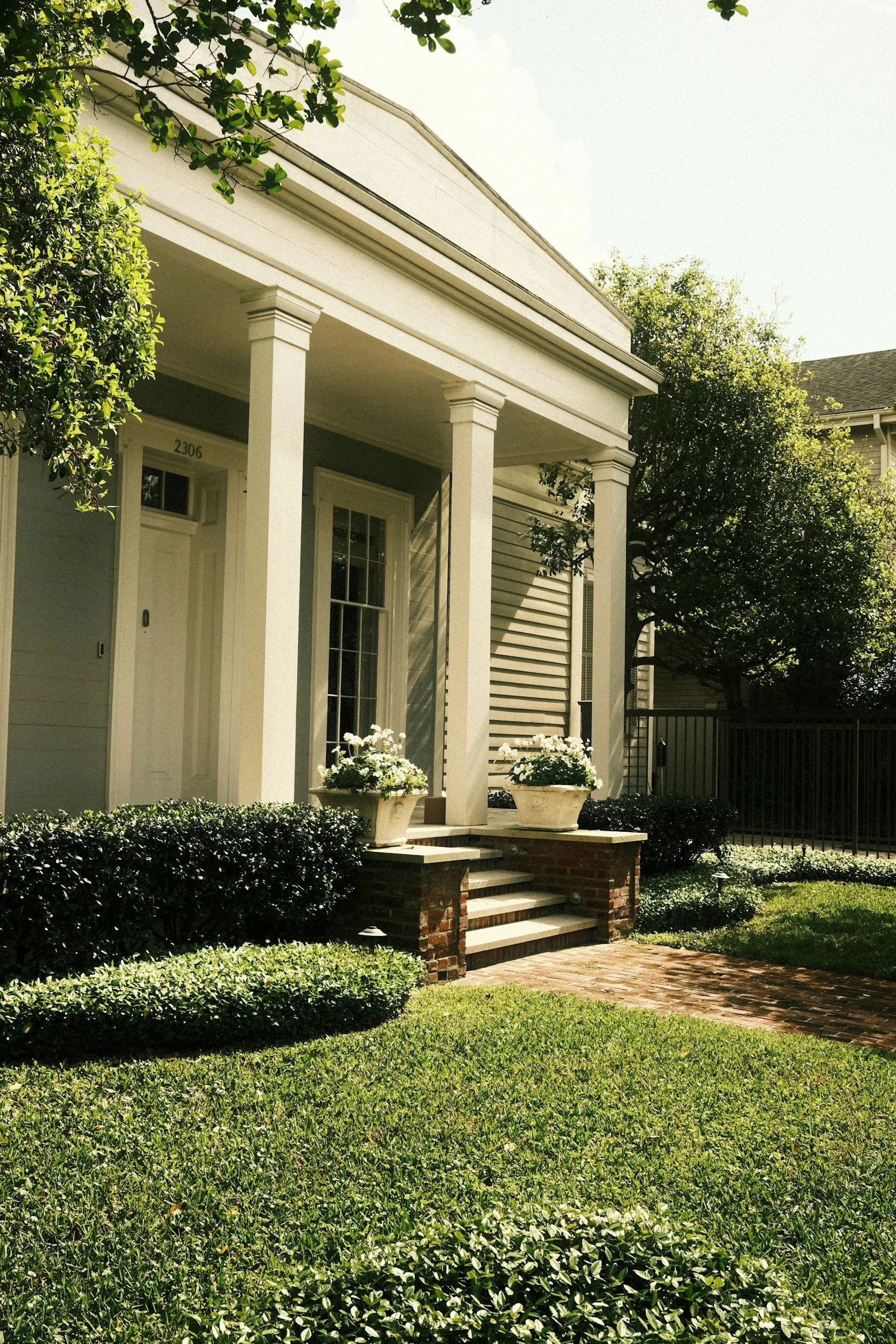 Front porch of a house with white columns, a brick staircase, and potted plants, surrounded by a well-maintained lawn and shrubs.
