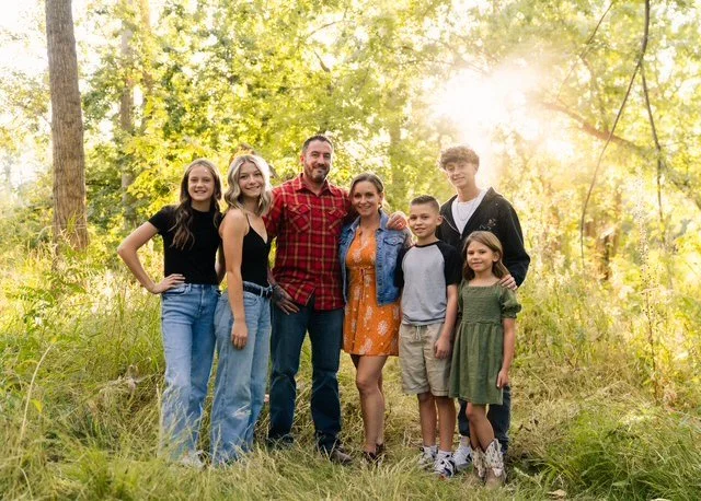 Family of seven posing outdoors in a forested area with sunlight filtering through trees.