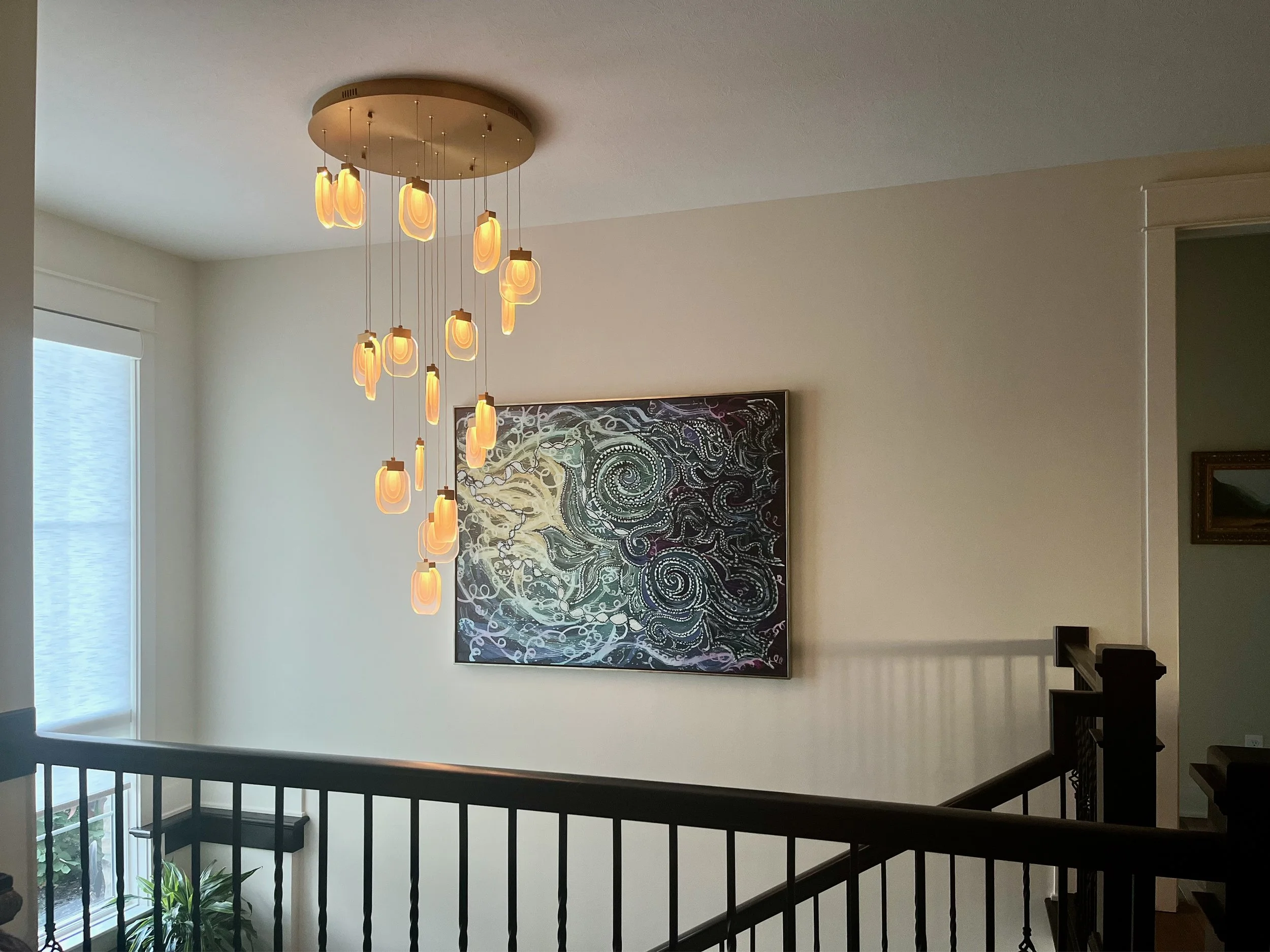 Interior view of a house showing a modern chandelier hanging from the ceiling, an abstract artwork on the wall, and a staircase railing in the foreground. A window provides natural light to the space.