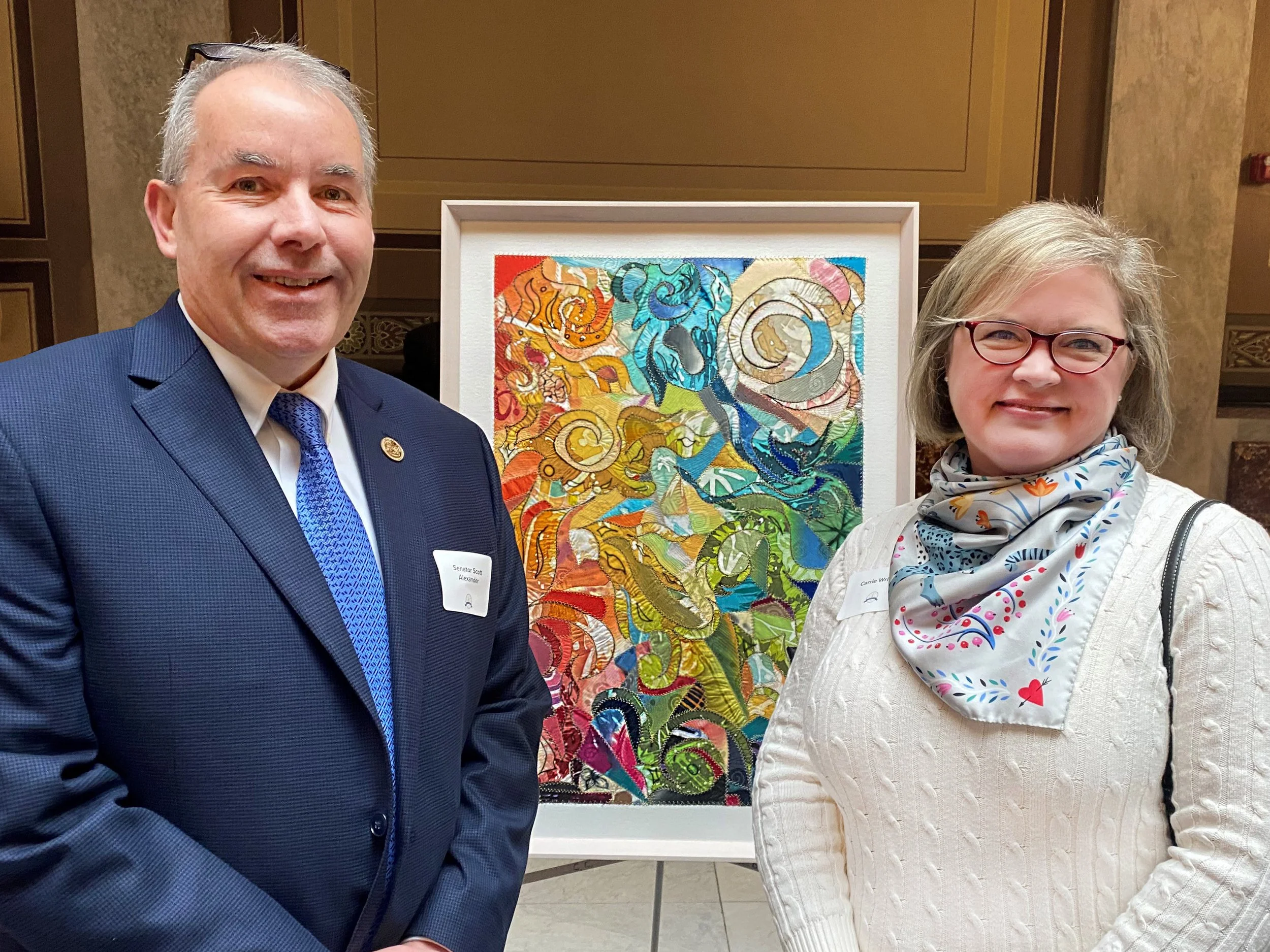 Two people standing in front of a colorful abstract artwork at an indoor event. The man on the left is wearing a dark blue suit, light dress shirt, and blue tie, with glasses resting on his head. The woman on the right is wearing glasses, a white textured sweater, and a white scarf with a colorful pattern. Both are smiling at the camera.