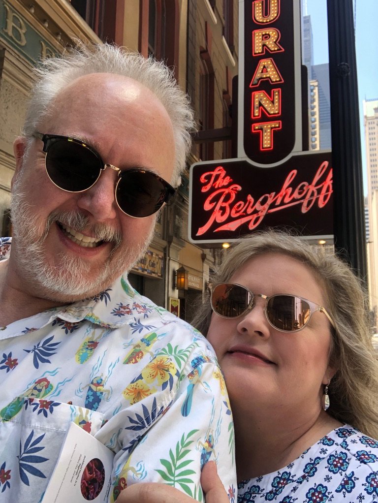 A smiling man and woman wearing sunglasses taking a selfie in front of a sign for The Bergdorf restaurant on a street in an urban area.
