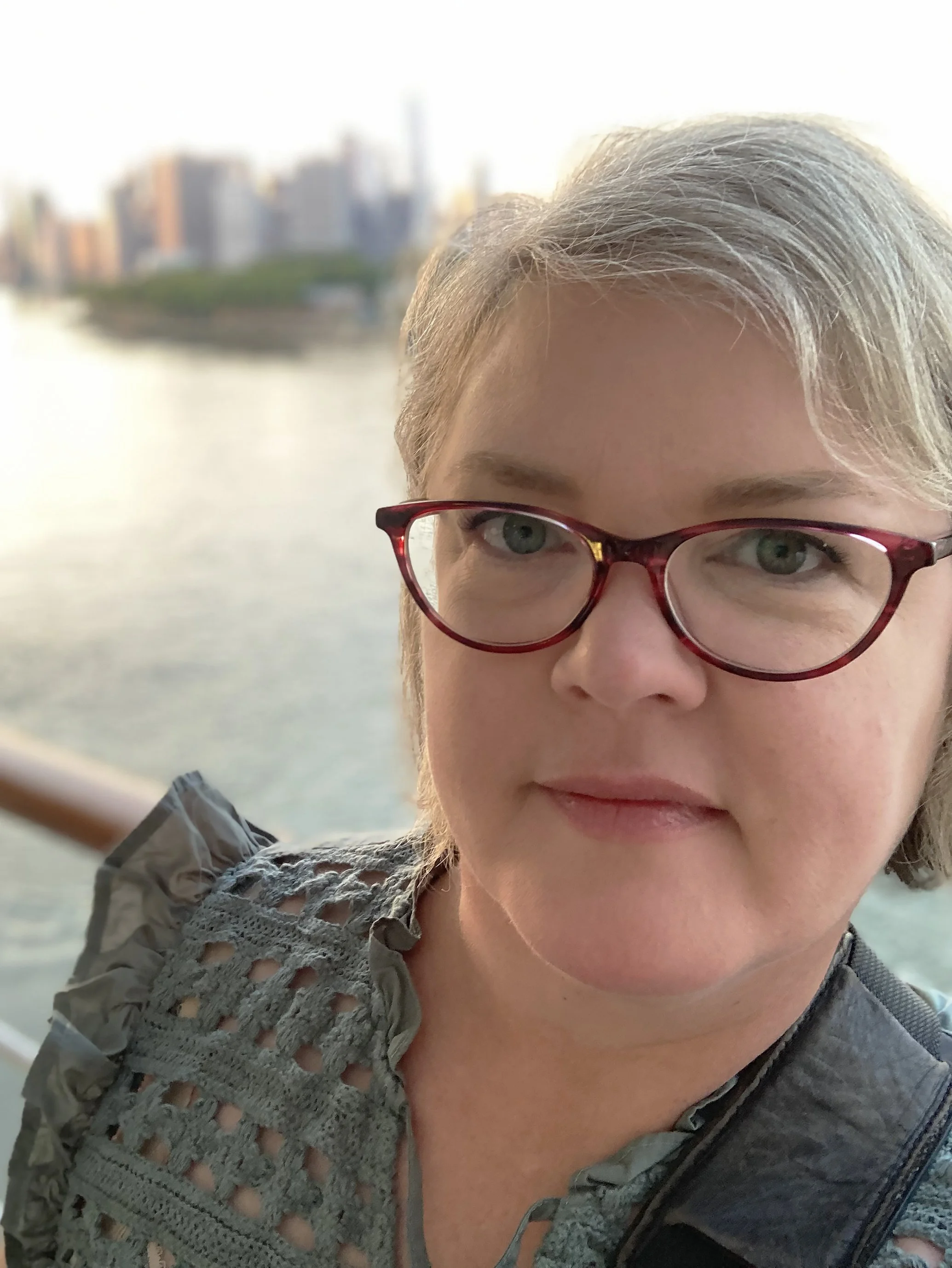 Close-up of a woman with gray hair and red glasses, smiling, with a city skyline across the water in the background.
