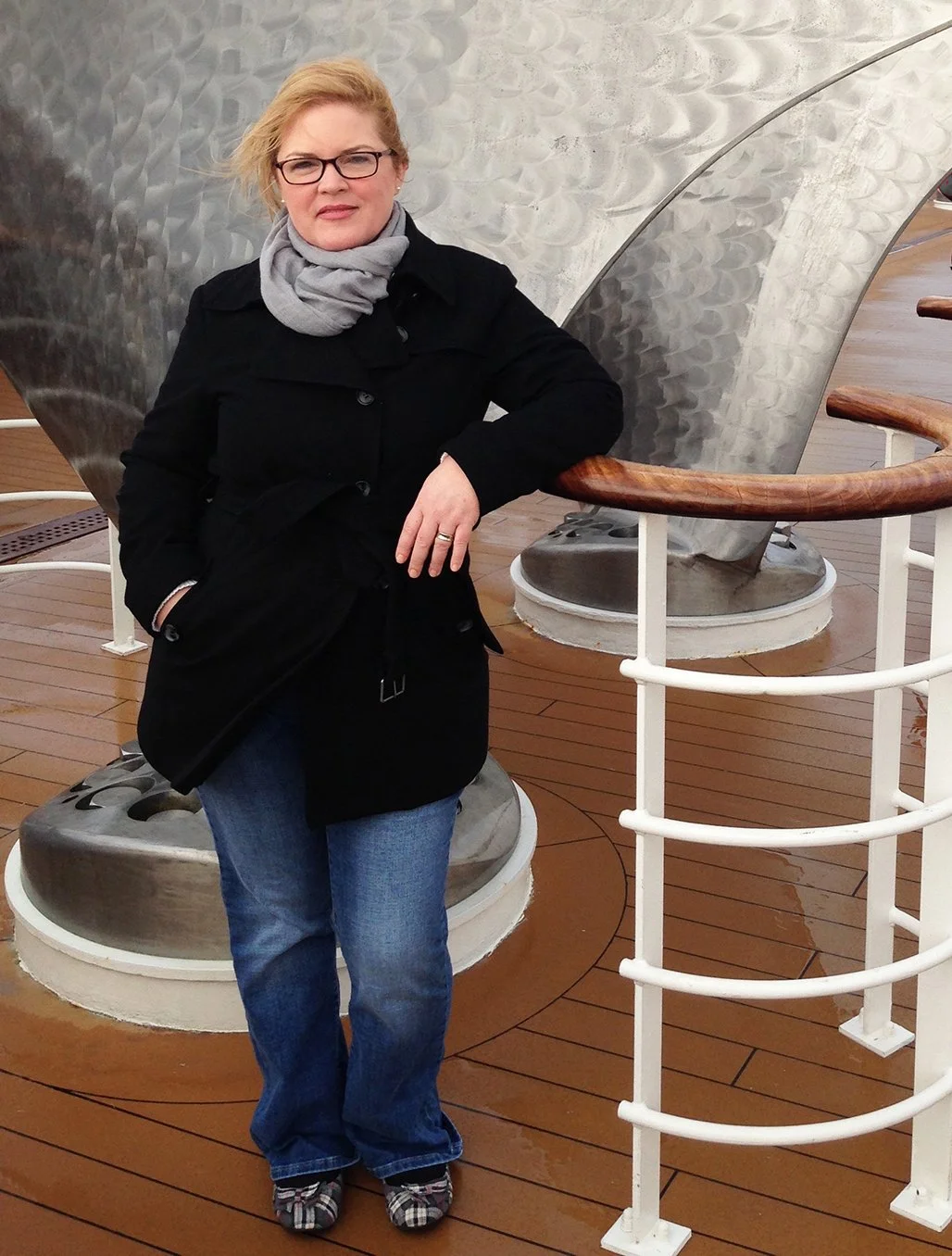 A woman with blonde hair, glasses, wearing a black coat, gray scarf, and blue jeans, standing on a wooden deck next to a large metallic ship structure.