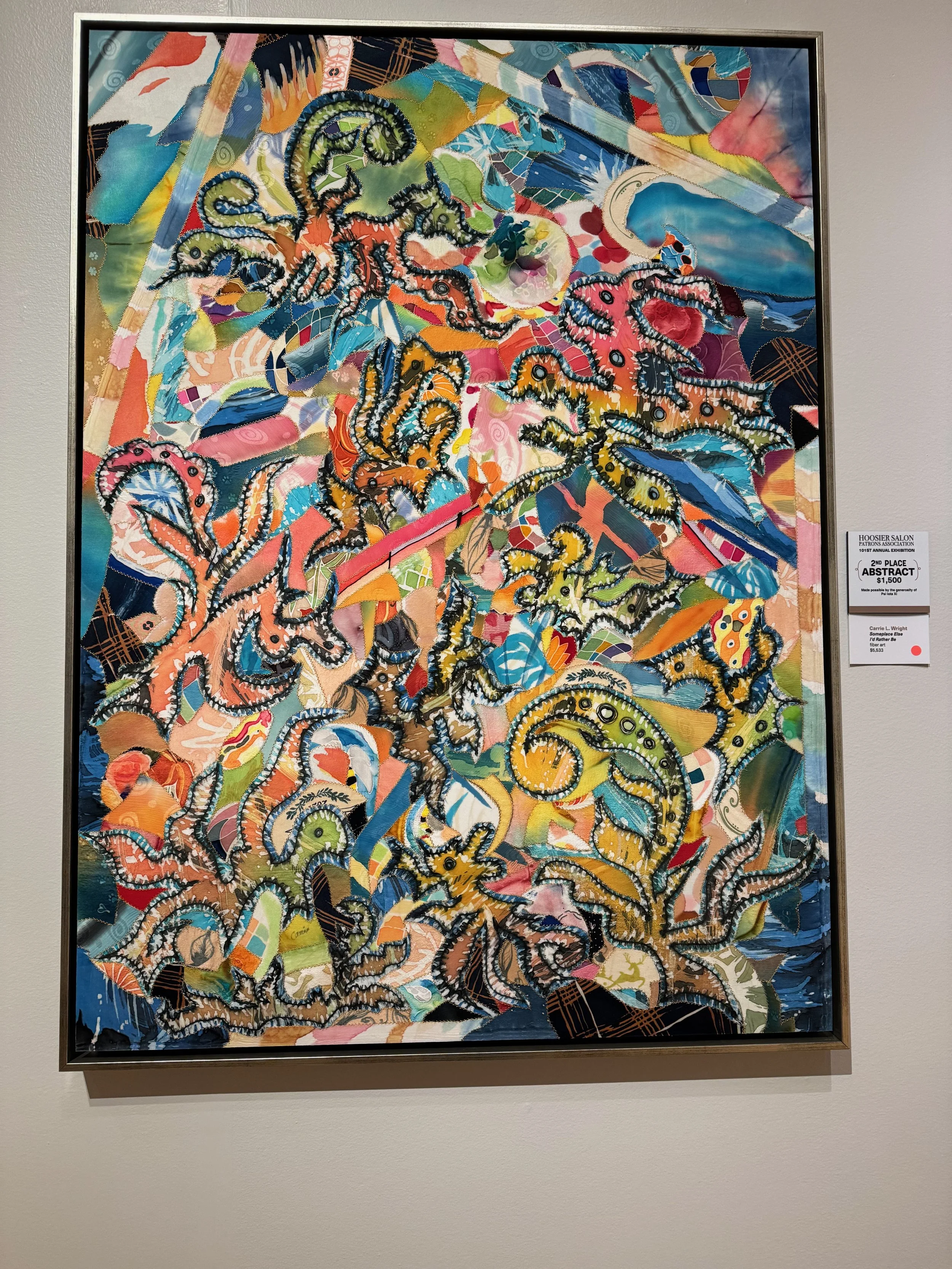 Colorful abstract quilted fabric artwork with various patterns and stitched designs, primarily featuring swirling lines and organic shapes, displayed in a silver frame on a white wall.