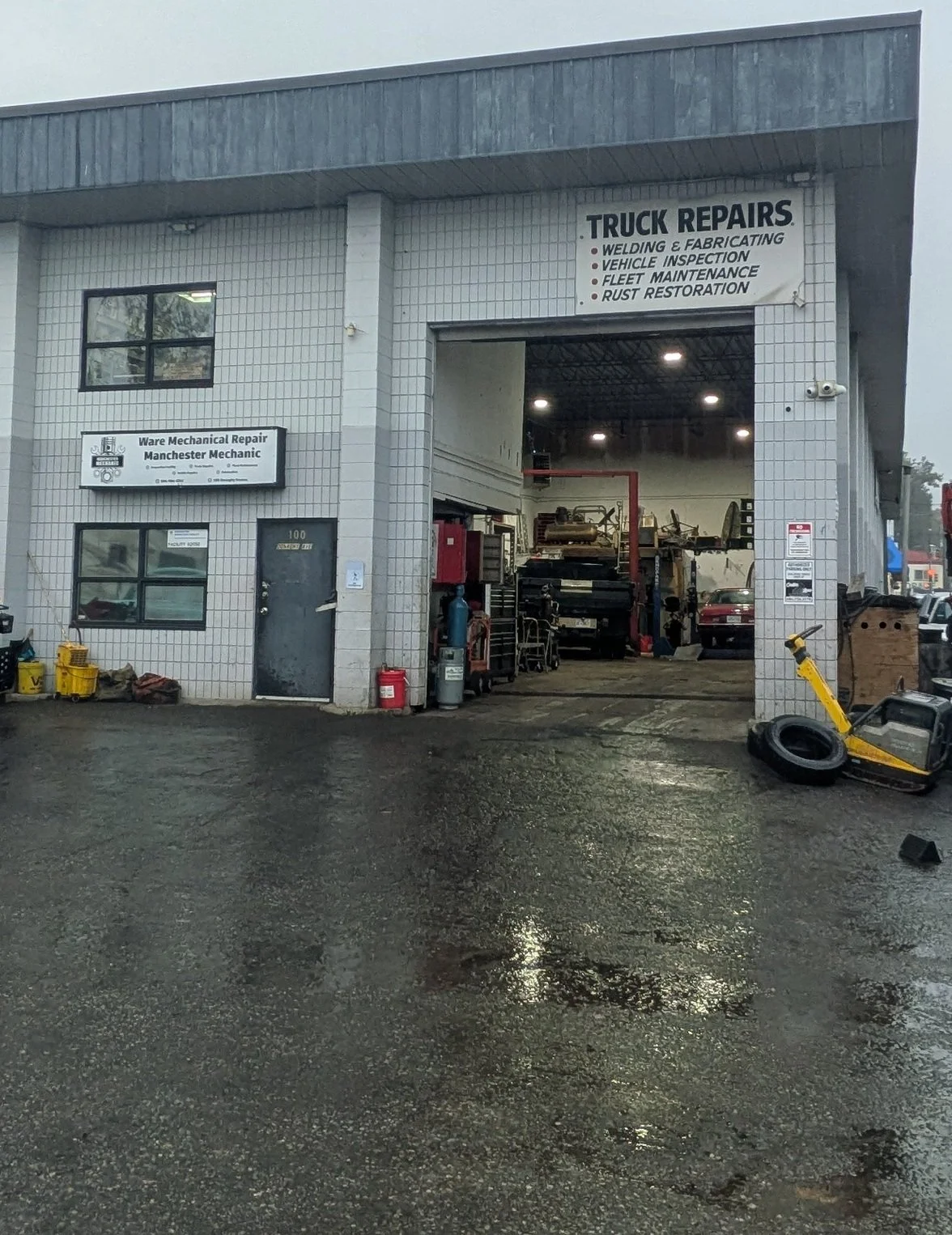 An auto repair shop with a large garage door opening to a workspace with vehicles and tools inside. Outside, there are yellow and black equipment, a red fire extinguisher, and a tire changer to the right, with a wet asphalt ground reflecting light.