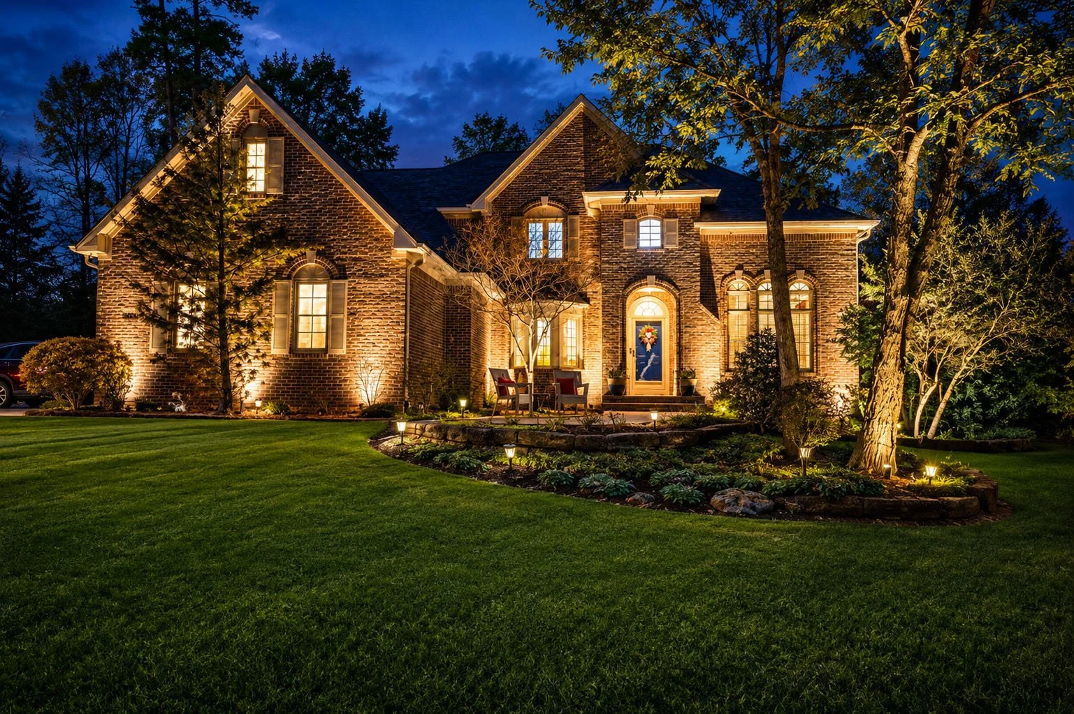 A brick house lit up at night with a well-manicured lawn and garden in the front yard, illuminated by outdoor lights.