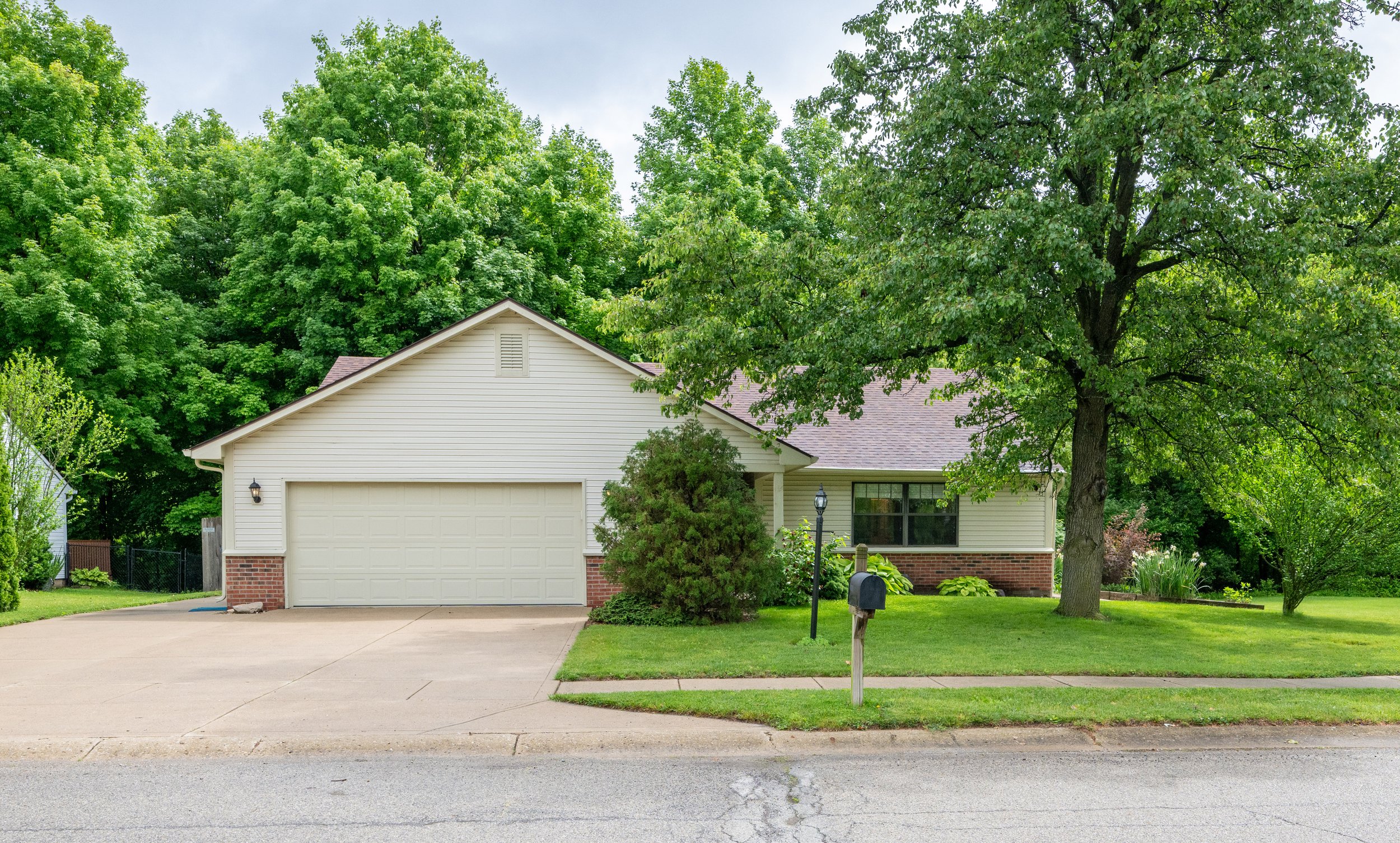 Front yard of a suburban house with a two-car garage, large green trees, a lawn, and a mailbox.