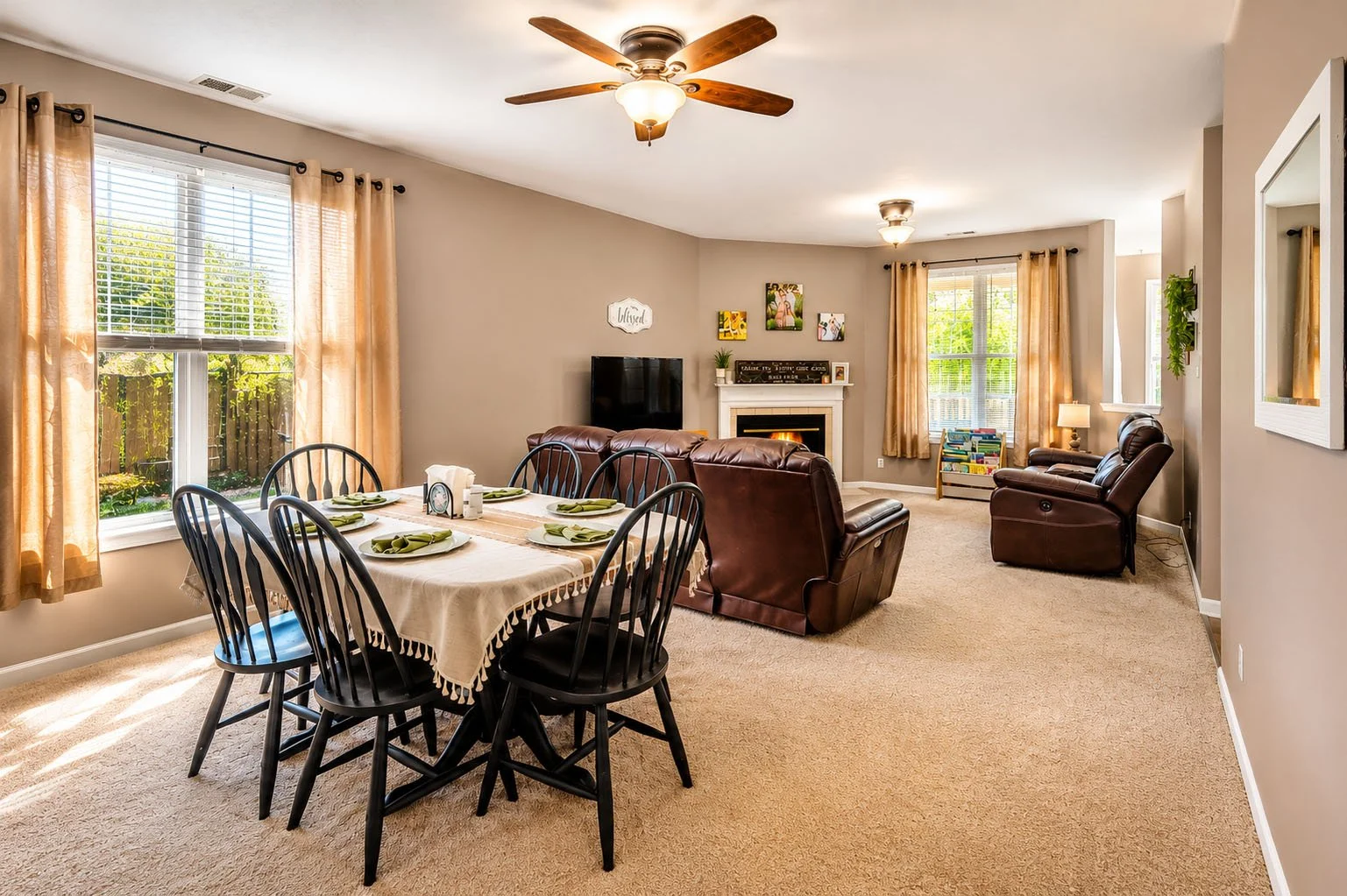 Living room and dining area with beige walls, tan carpet, black dining table with six chairs, brown leather sofas, a fireplace, a flat-screen TV, and windows with beige curtains letting in sunlight.