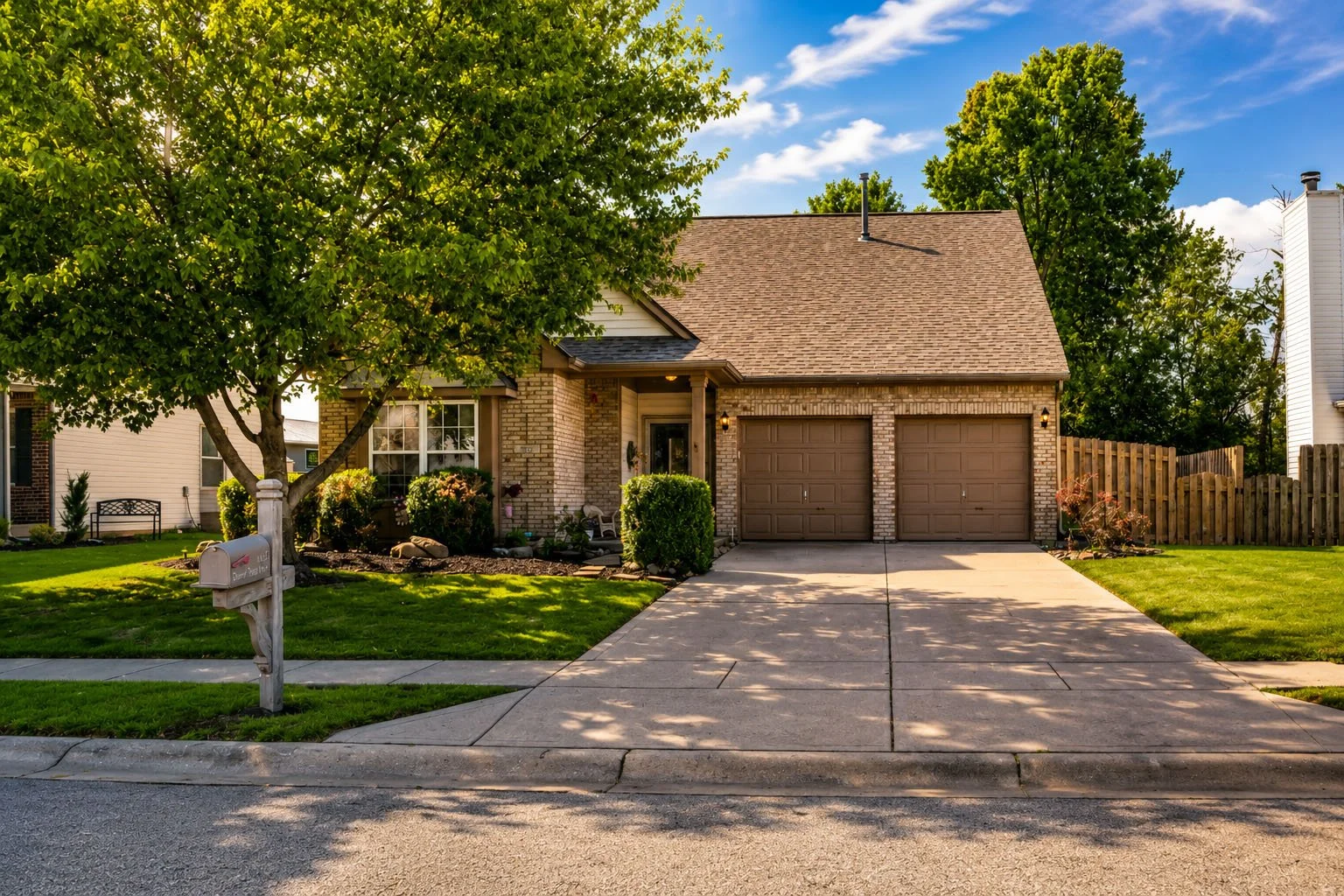 A suburban house with a brick and beige exterior, two-car garage, front porch, and well-maintained green lawn with a tree and shrubs.