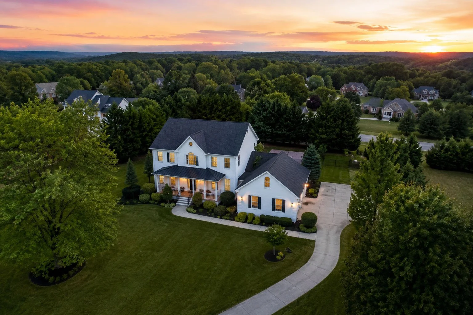 A two-story white house with black shutters, surrounded by a well-maintained lawn and trees, during sunset in a suburban neighborhood.
