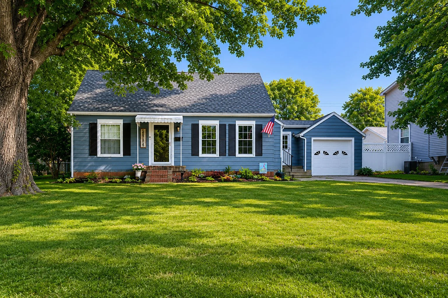 A blue suburban house with black shutters, white trim, and a small front porch with brick steps. It has a white garage door, American flag, and a well-maintained lawn with trees and plants.