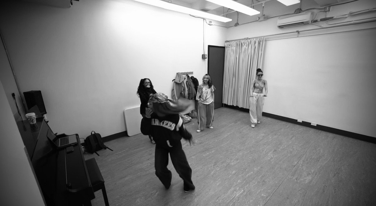 Four women in a dance studio with a plain wall, curtain, and coat rack, practicing or rehearsing dance moves.