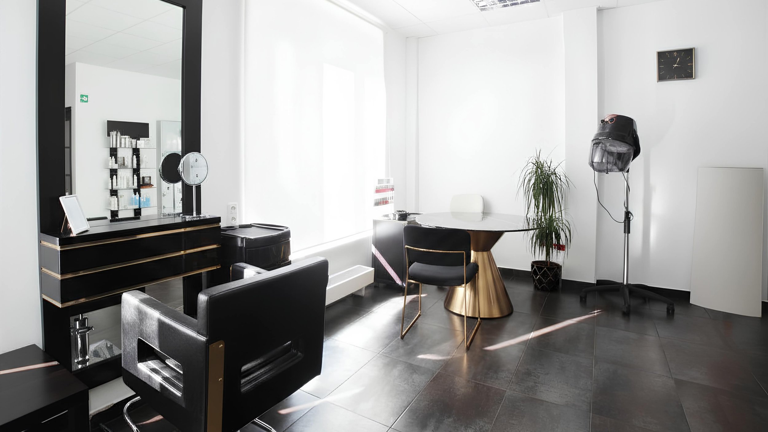 A modern hair salon with black and gold furniture, a large mirror, a hair salon chair, a viewing station, a table with a plant, a hair dryer, and a clock on the wall, illuminated by natural light.