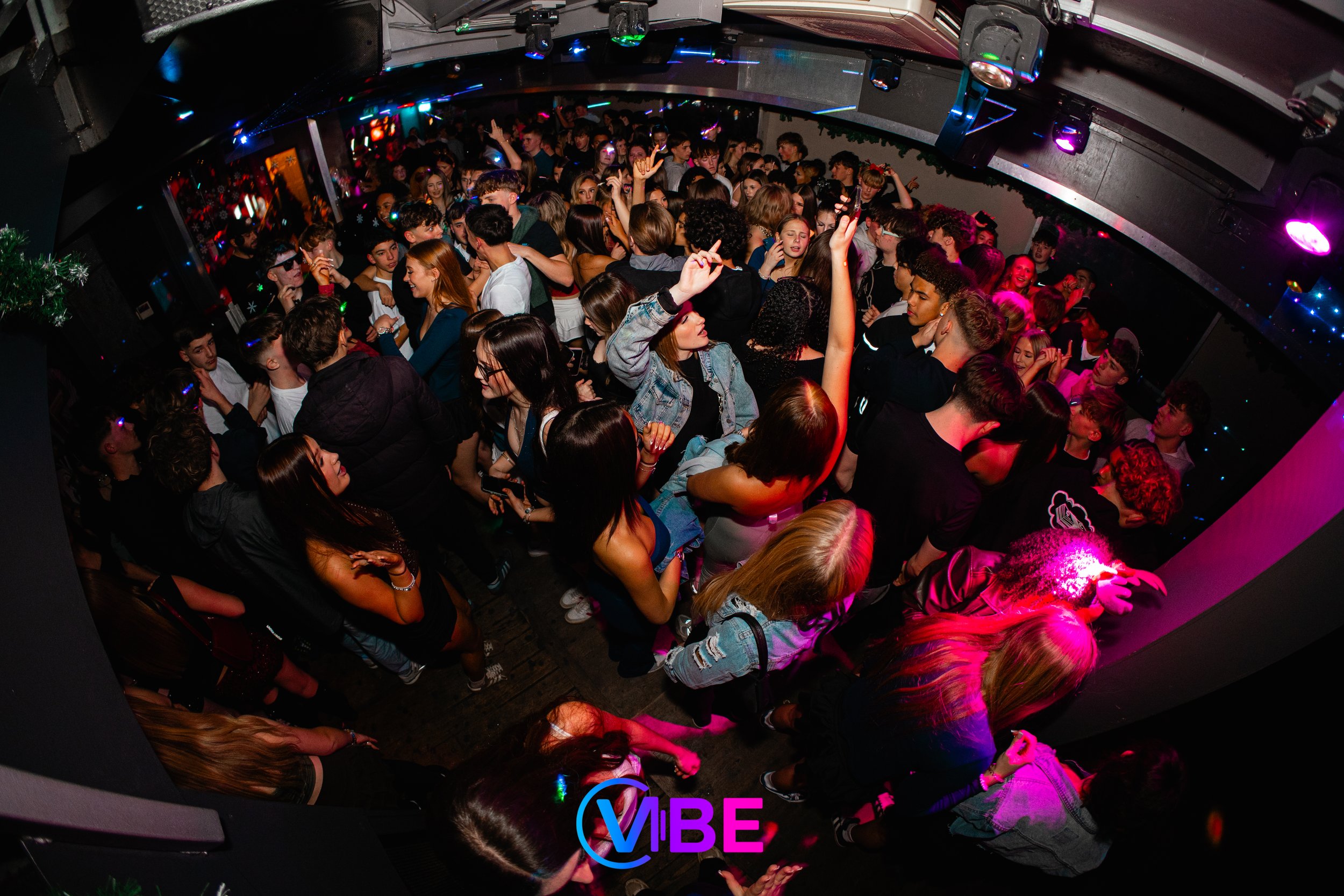 A crowded dance floor filled with young people enjoying a night out at a club, with colorful lights and a neon sign that says 'VIBE'.