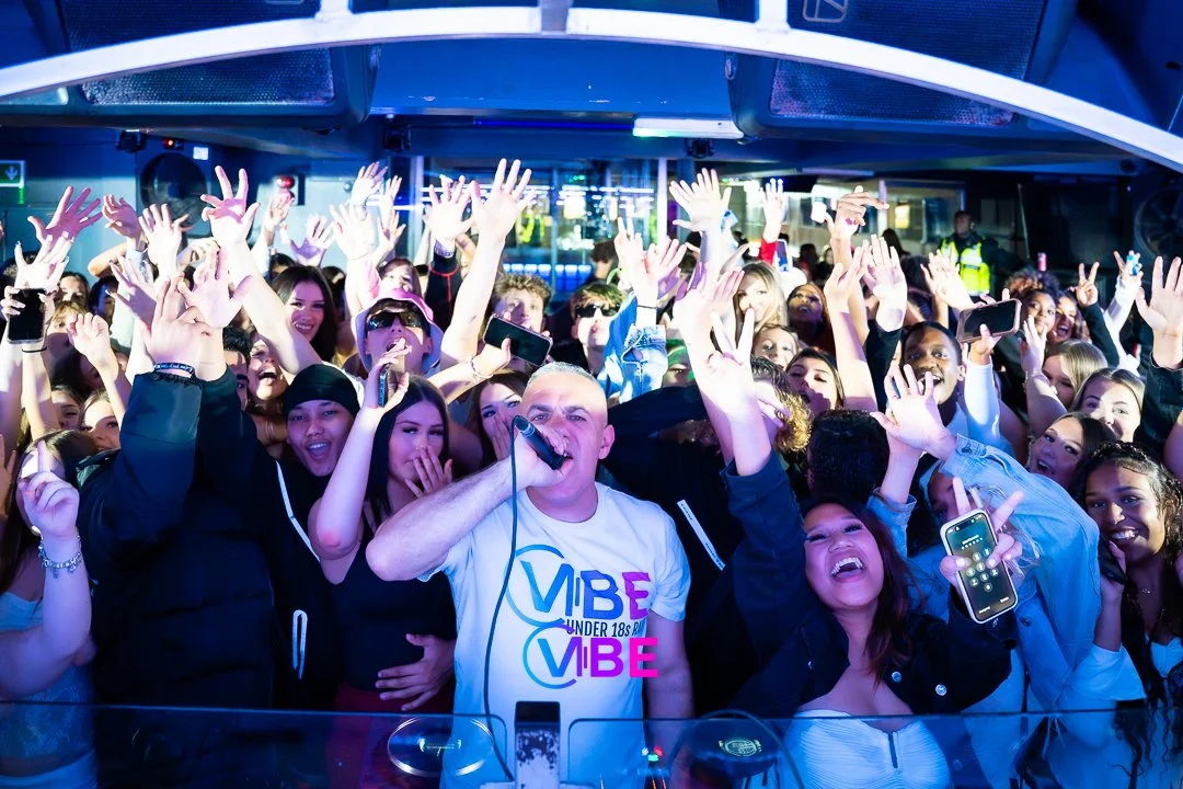 Crowd of young people dancing and celebrating at a party or concert, with a male DJ in the foreground holding a microphone.
