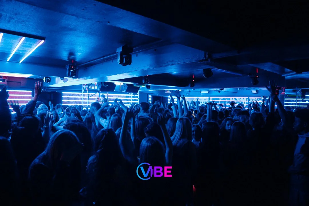 Crowd of people dancing and raising their hands in a nightclub with blue lighting and horizontal light bars.