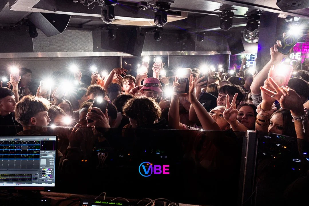 Crowd of people inside a nightclub or concert venue, illuminated by numerous smartphone flashlights, with DJ equipment and a colorful 'VIBE' sign in the foreground.