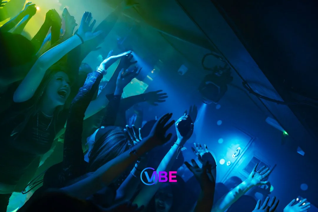 People dancing and enjoying at a nightclub with colorful lights and the word "VIBE" displayed on the screen.