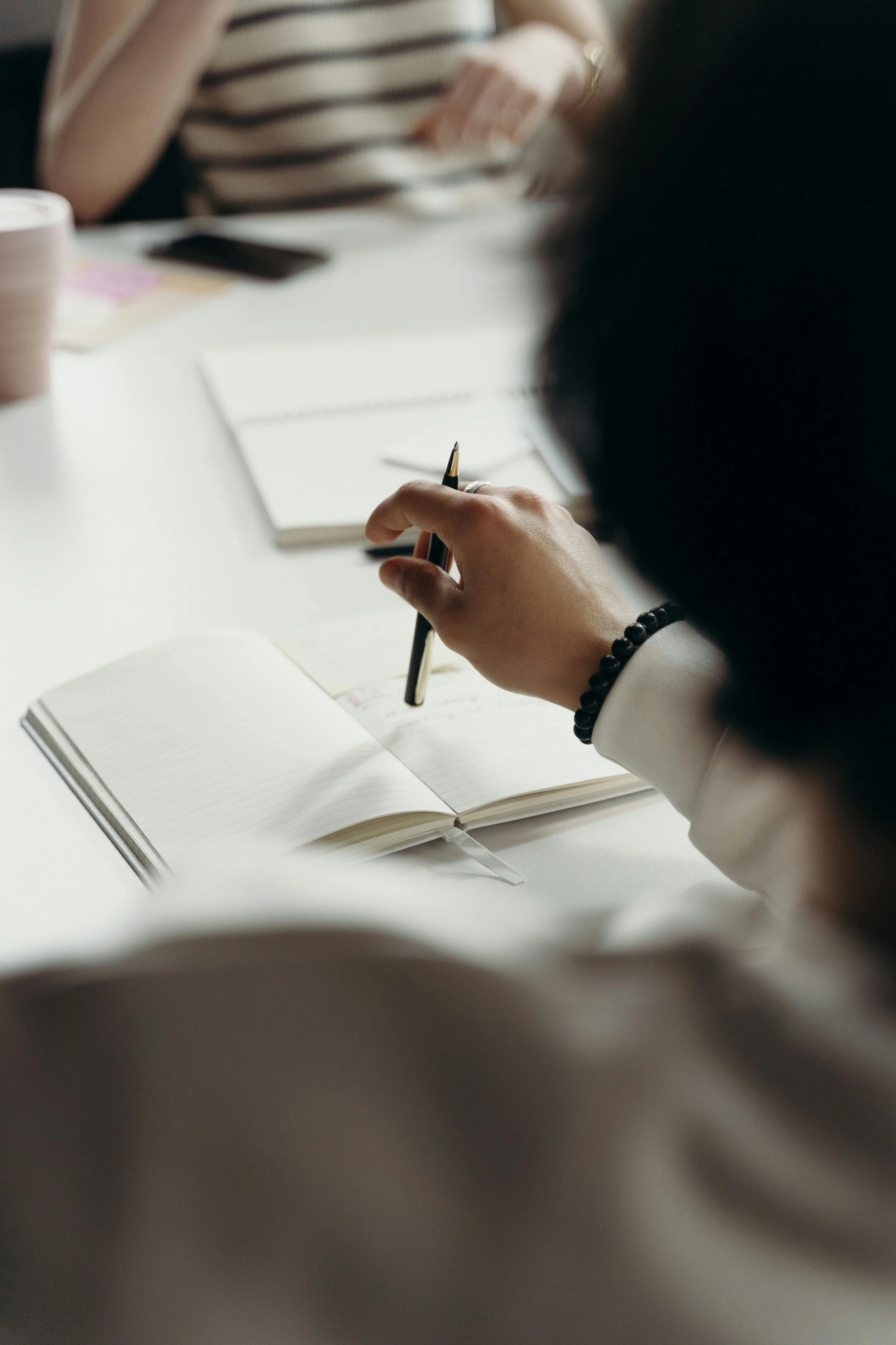 Close-up of a person holding a pen and writing in an open notebook on a white surface, with other writing materials and a person in a striped shirt visible in the blurry background.