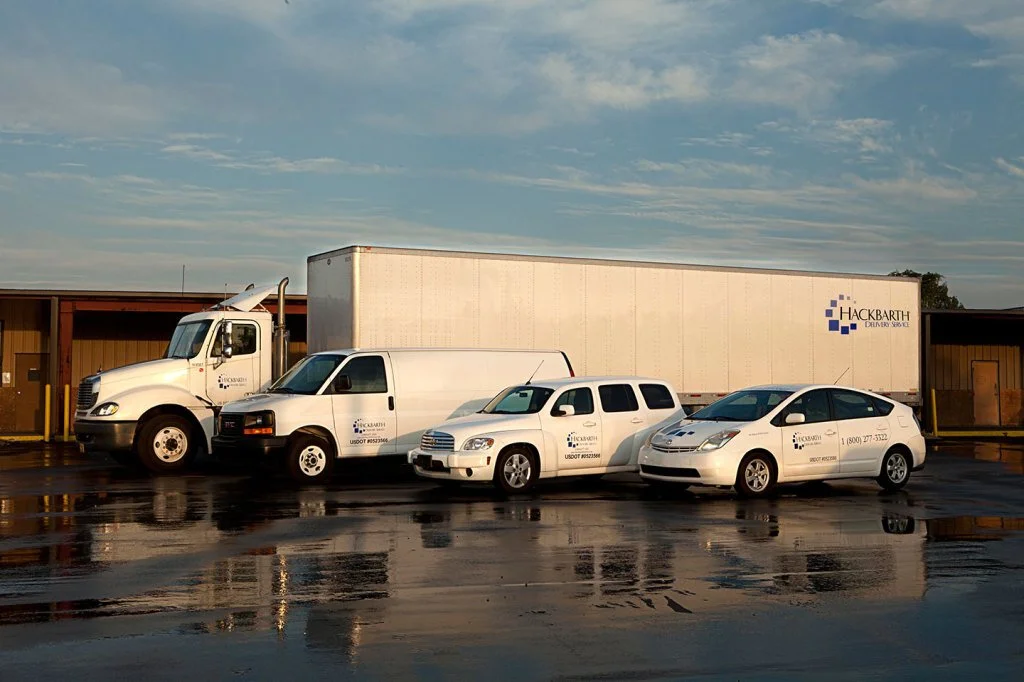 Four vehicles, including a large delivery truck, a white van, a white SUV, and a white car, parked side by side near a building with a cloudy sky overhead.