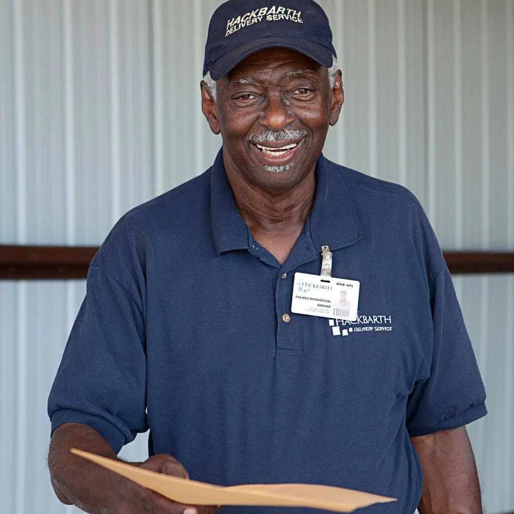 An older man smiling, wearing a navy blue cap and shirt with Hackbarth Delivery Service logos, holding a yellow envelope, standing indoors.