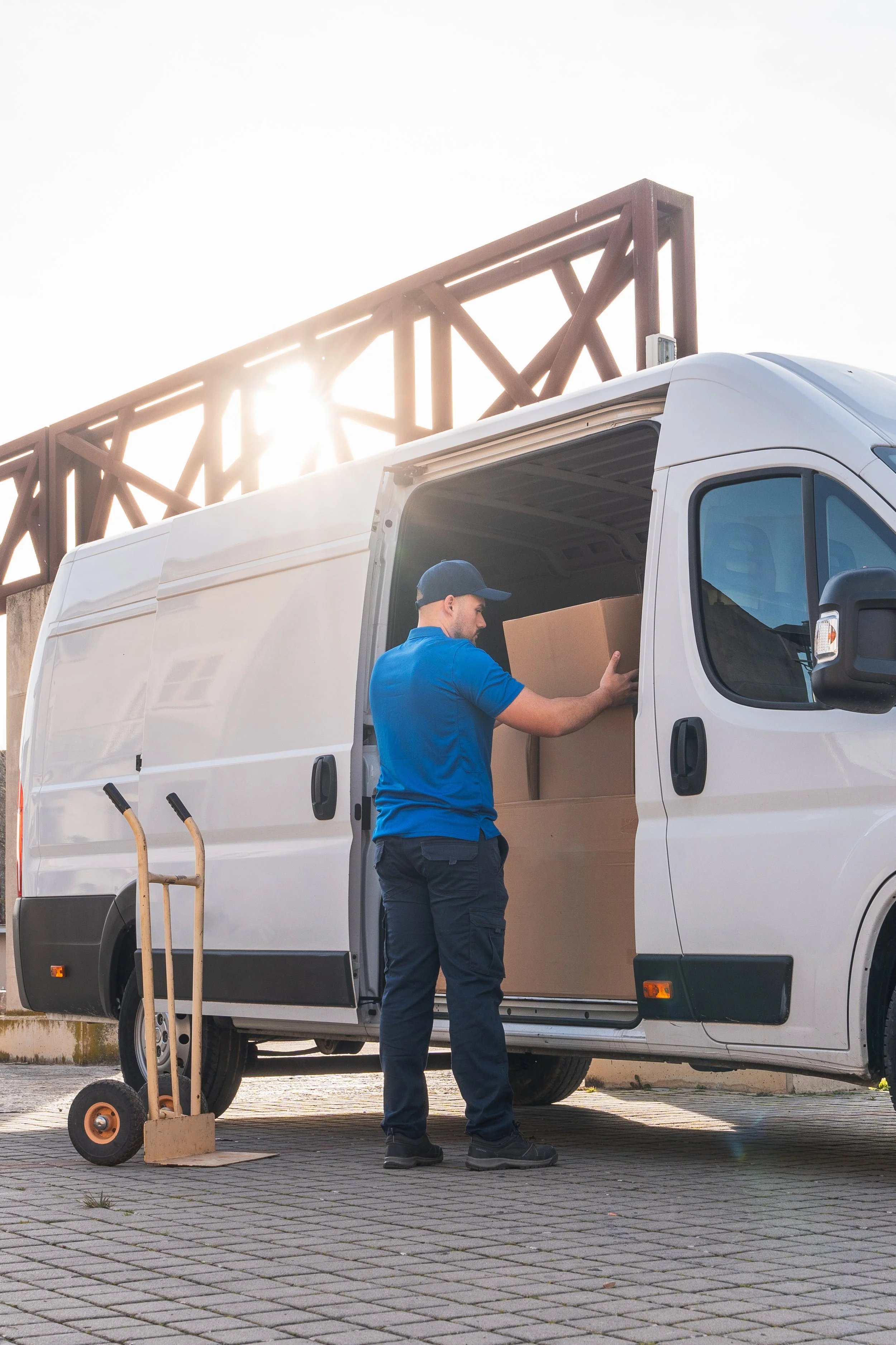 A delivery person wearing a blue uniform and black boots loading packages into a white delivery van. A hand truck with a cardboard box and a dolly are nearby, set against a brick sidewalk with a wooden fence in the background.