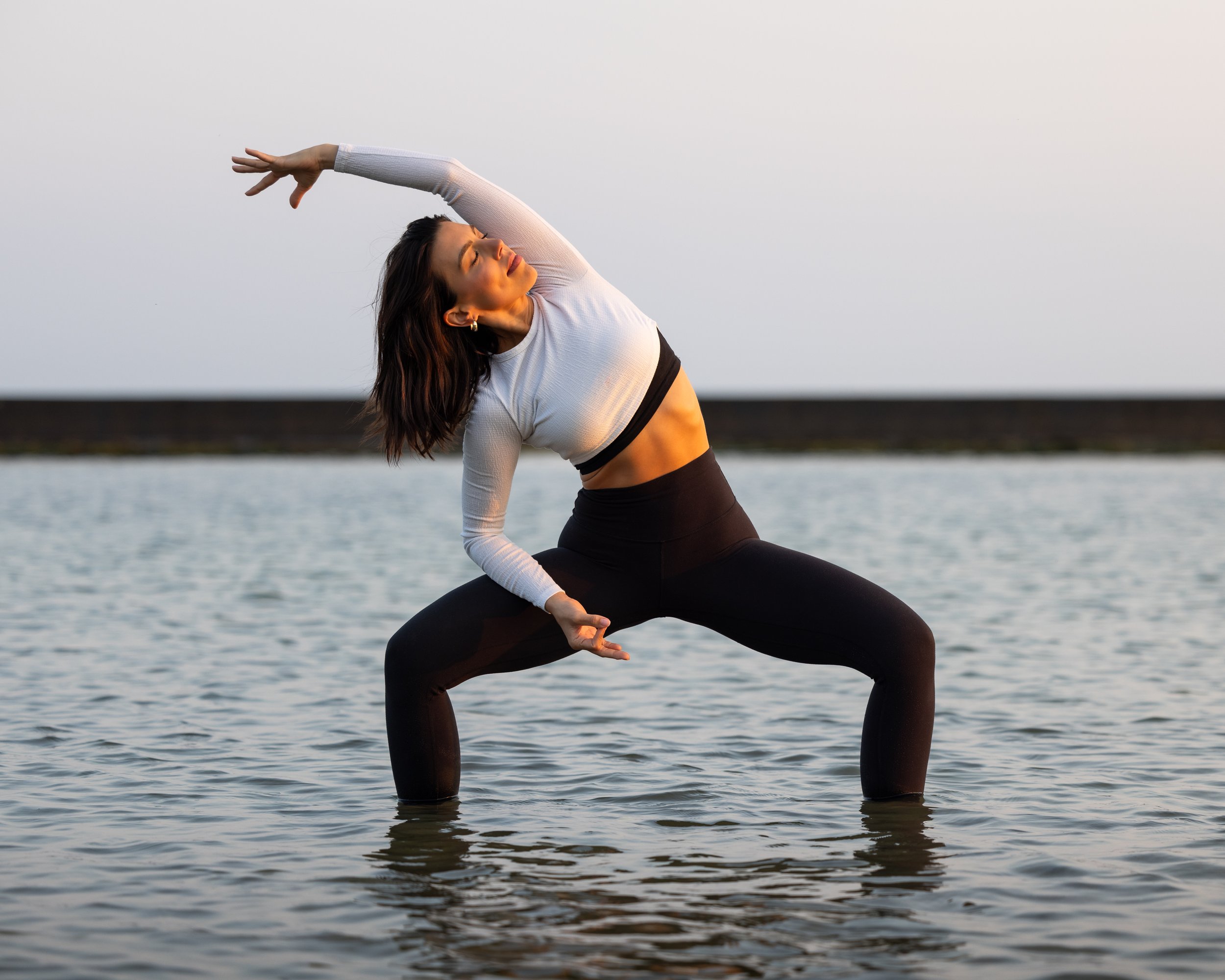 A woman in black leggings and a white long-sleeve top practicing yoga in shallow water at sunset, with her eyes closed and arms raised in a side bend pose.