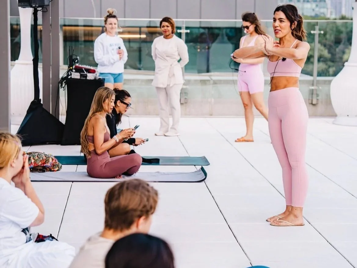 A woman in a pink workout outfit leading a fitness or yoga class outdoors, with people sitting and standing around her, some taking photos or listening attentively.