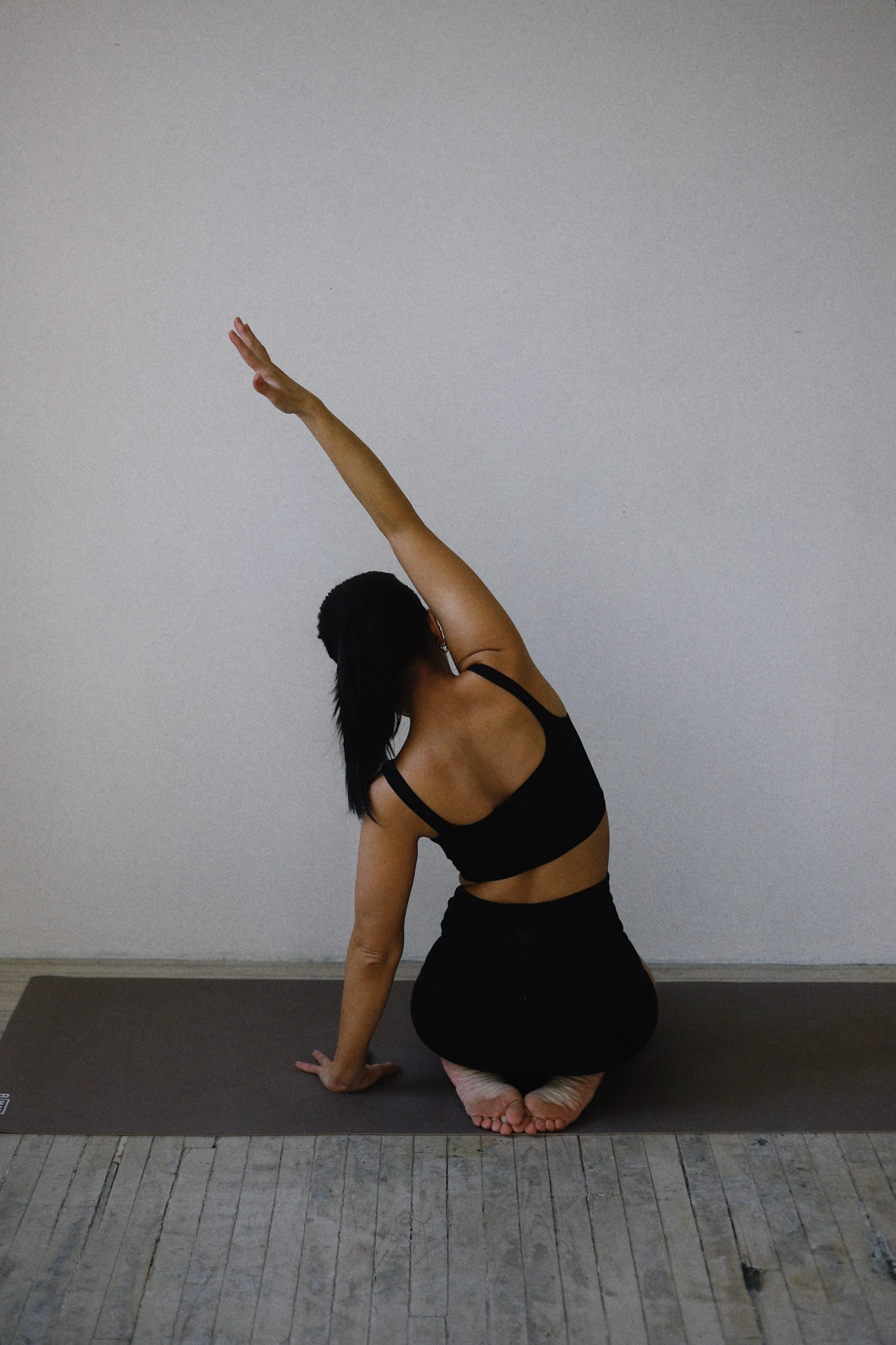 A woman practicing yoga indoors, kneeling on a yoga mat with one hand on the ground and the other reaching upward, against a plain white wall.