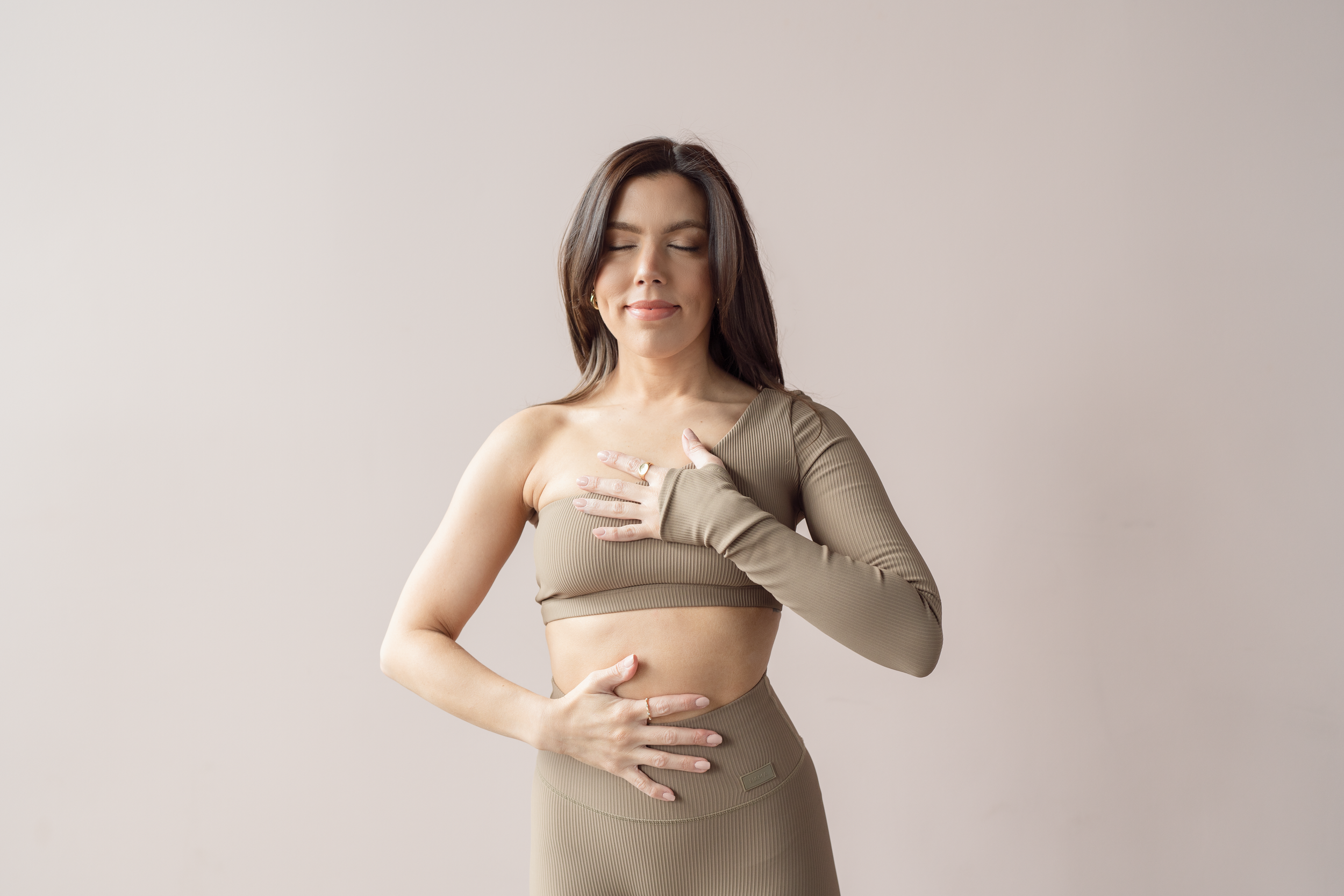 A woman with long dark hair and closed eyes is standing against a plain beige background. She is wearing a beige, one-shoulder, long-sleeve top and matching high-waisted bottom. She has one hand on her chest and the other on her stomach, with a gentle smile on her face.