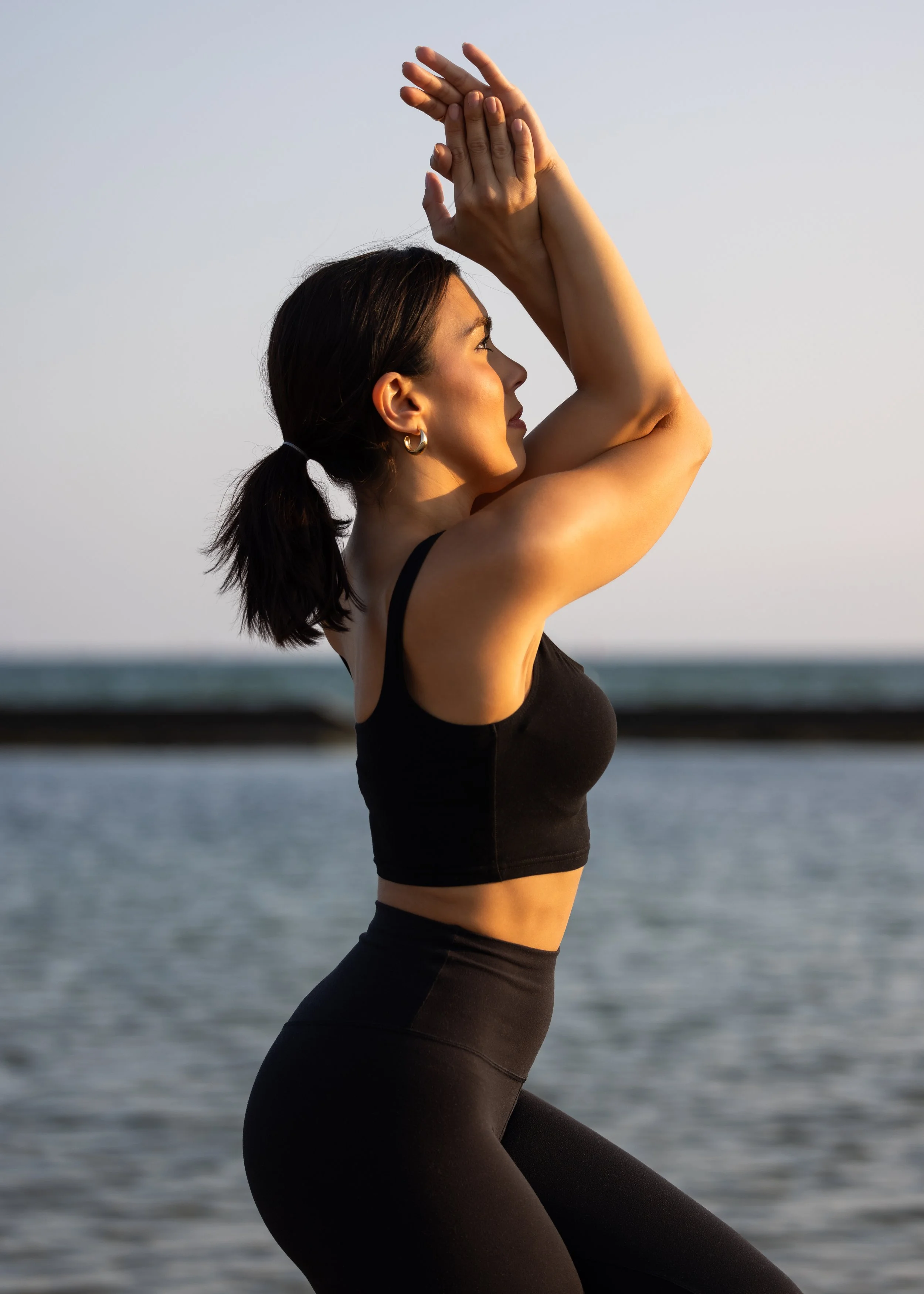 A woman practicing yoga near the beach with her arms raised above her head, wearing a black tank top and leggings, with the ocean and sky in the background during sunset.