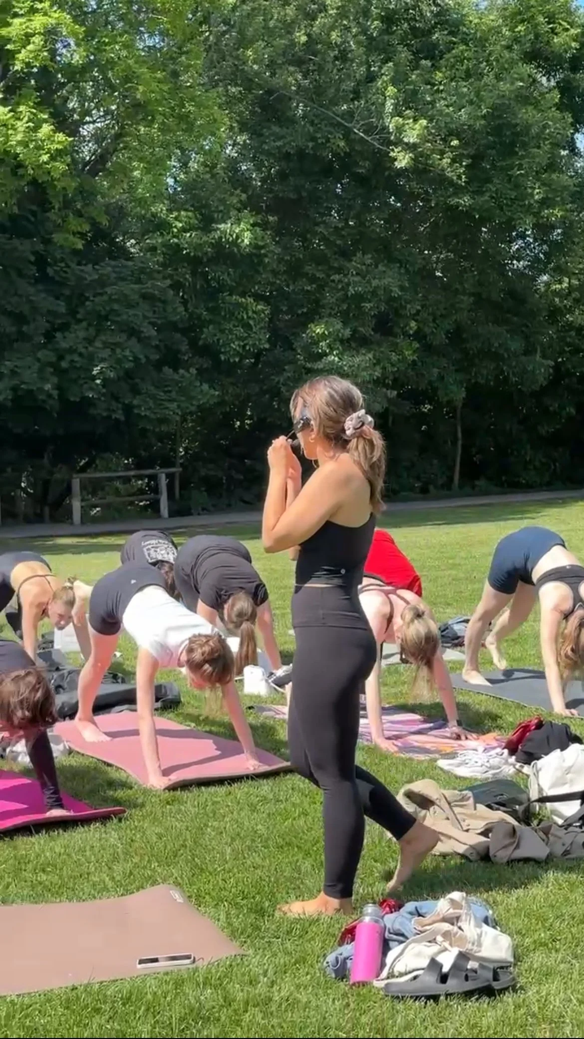 A woman in black leggings and a black tank top standing outdoors during a yoga class with others bent over on yoga mats on grass, surrounded by trees.