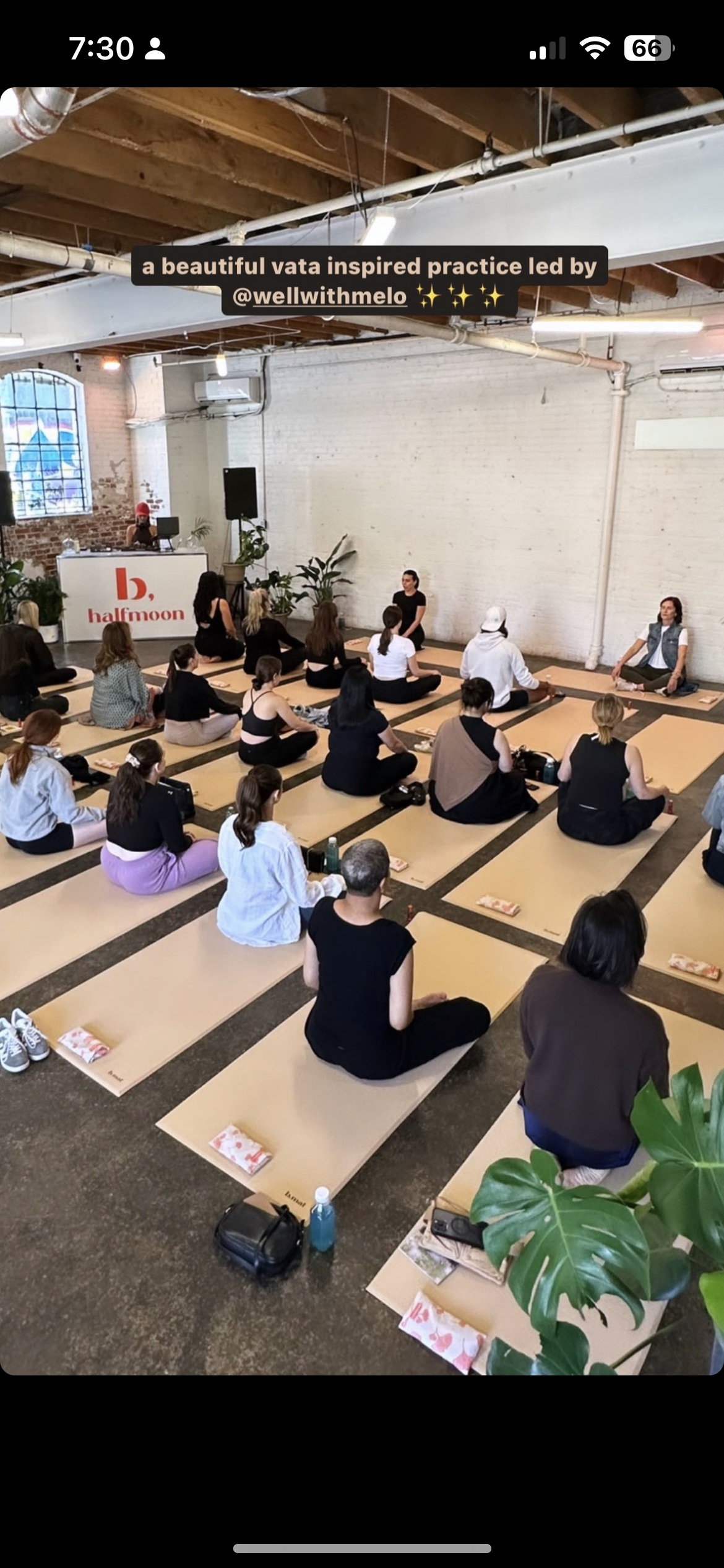 People practicing yoga in a studio with wooden beams, exposed brick wall, and large windows, sitting on mats in a group class led by an instructor. The studio has plants and a DJ setup at the front.