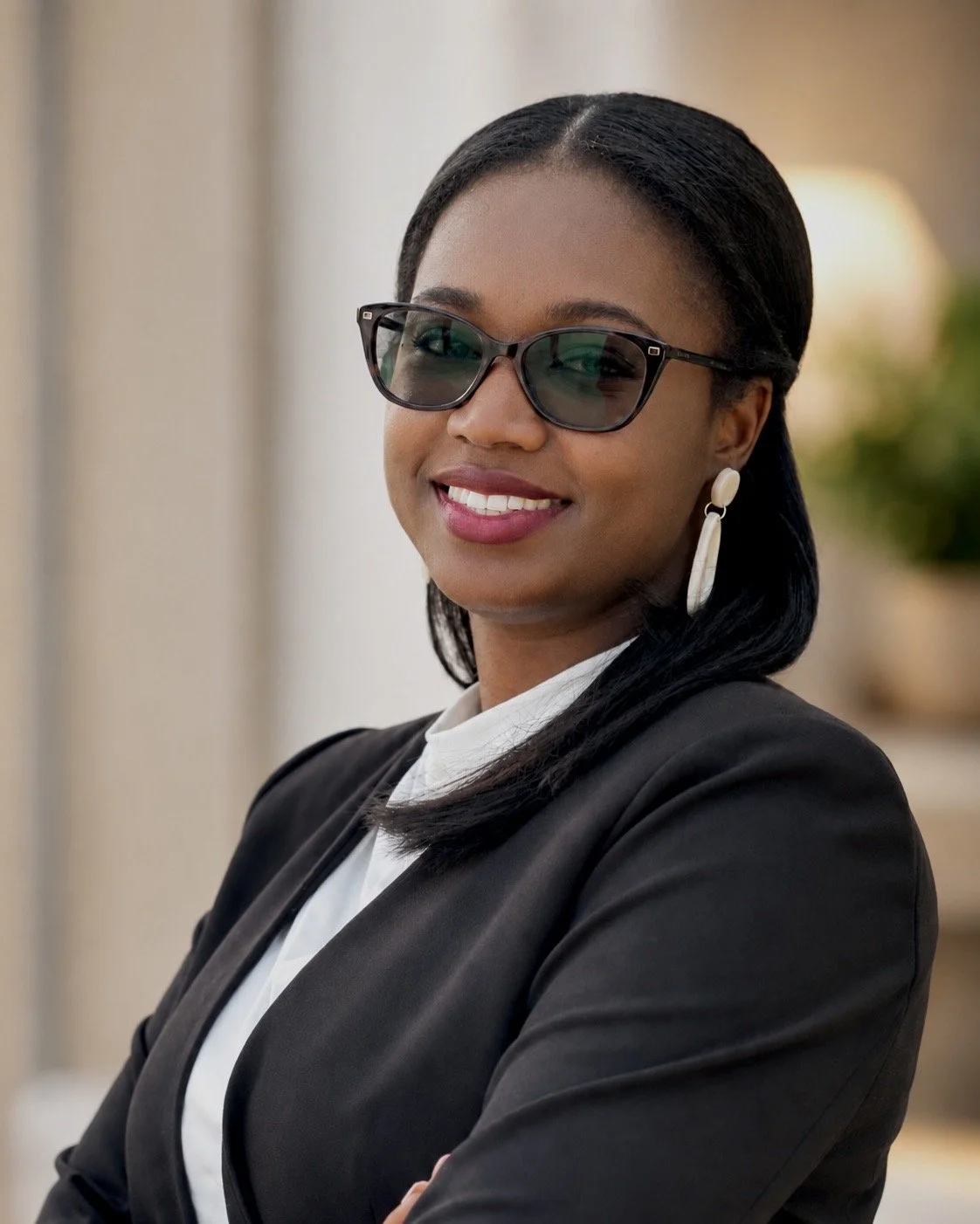 A professional woman with dark hair, wearing glasses, earrings, a black blazer, and a white shirt, smiling confidently indoors.