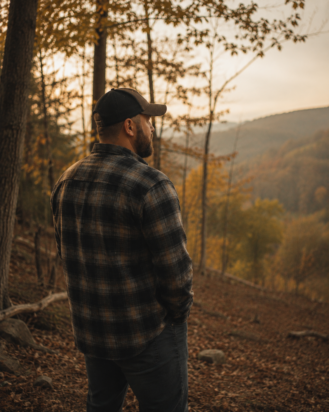 A man in a plaid shirt and baseball cap standing in a wooded area during autumn, looking at the mountains in the distance.