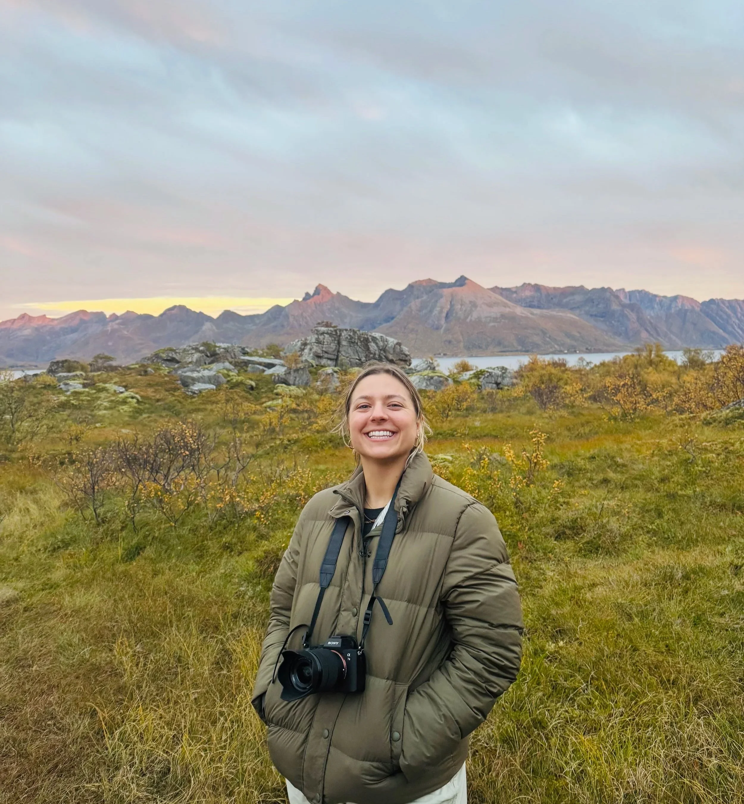 A woman smiling outdoors in a grassy field with mountains in the background and a cloudy sky.