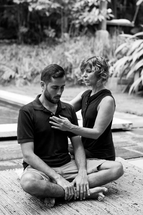 A woman is performing a spiritual or healing energy practice on a seated man outdoors near a body of water.
