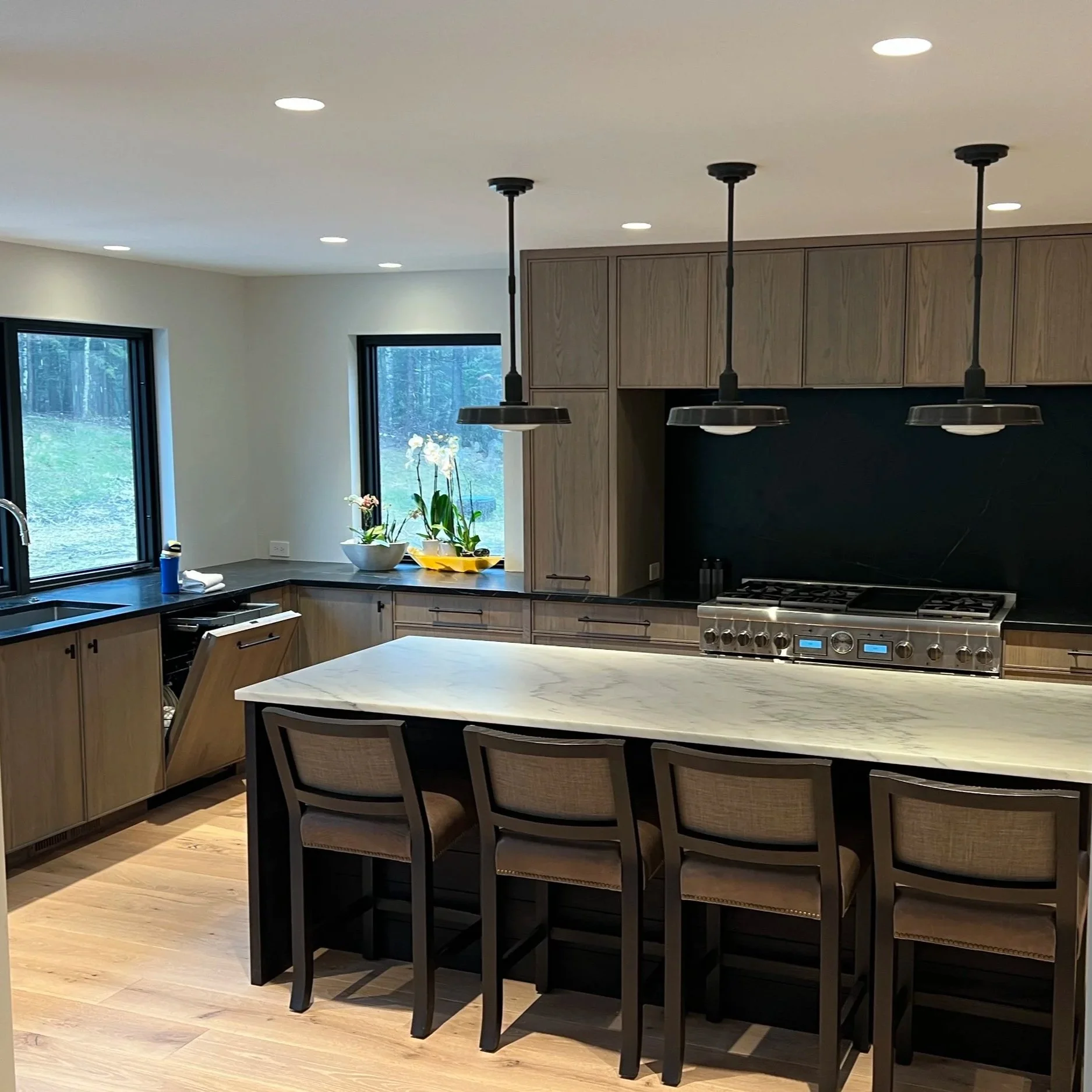 Modern kitchen with light wooden cabinets, black countertops, black kitchen island with chairs, stainless steel stove, hanging pendant lights, and large windows overlooking a green outdoor area.