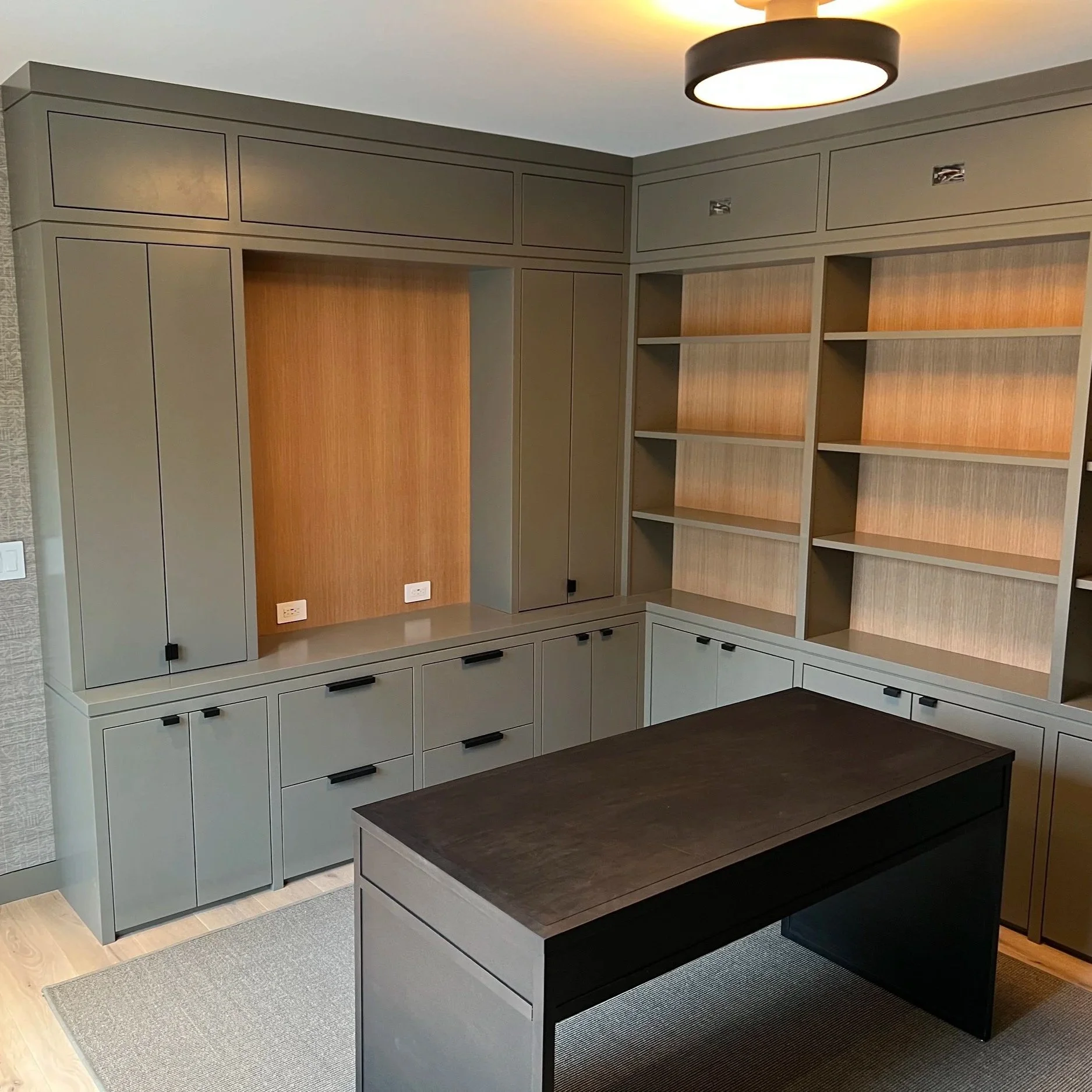 Modern built-in shelving and cabinetry unit in neutral tones with black handles, a dark wood table, gray and beige area rugs, ceiling light fixture, and electrical outlets, in a contemporary interior space.